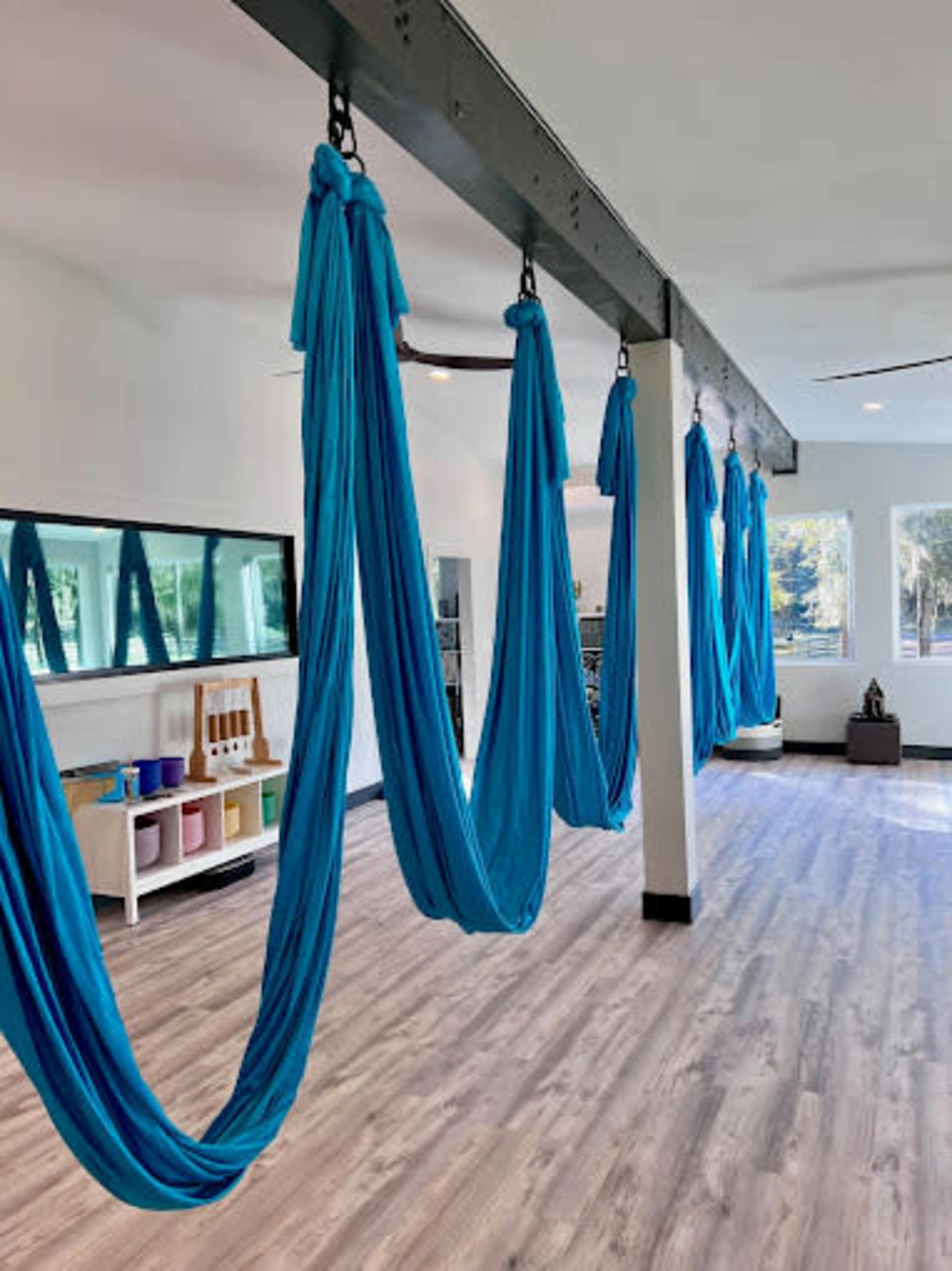 The image shows an indoor room equipped with blue aerial silks hanging from a wooden beam, with a mirrored wall and light-colored flooring.