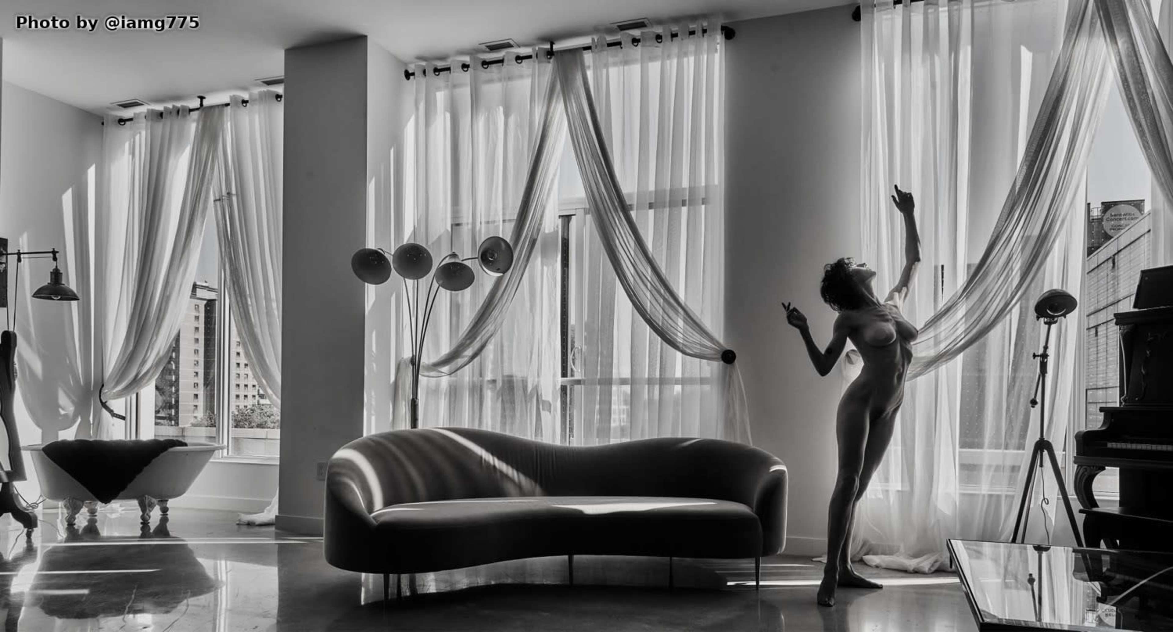 A figure stands gracefully in a sunlit room with large windows draped in flowing curtains, beside a curved sofa and a piano.