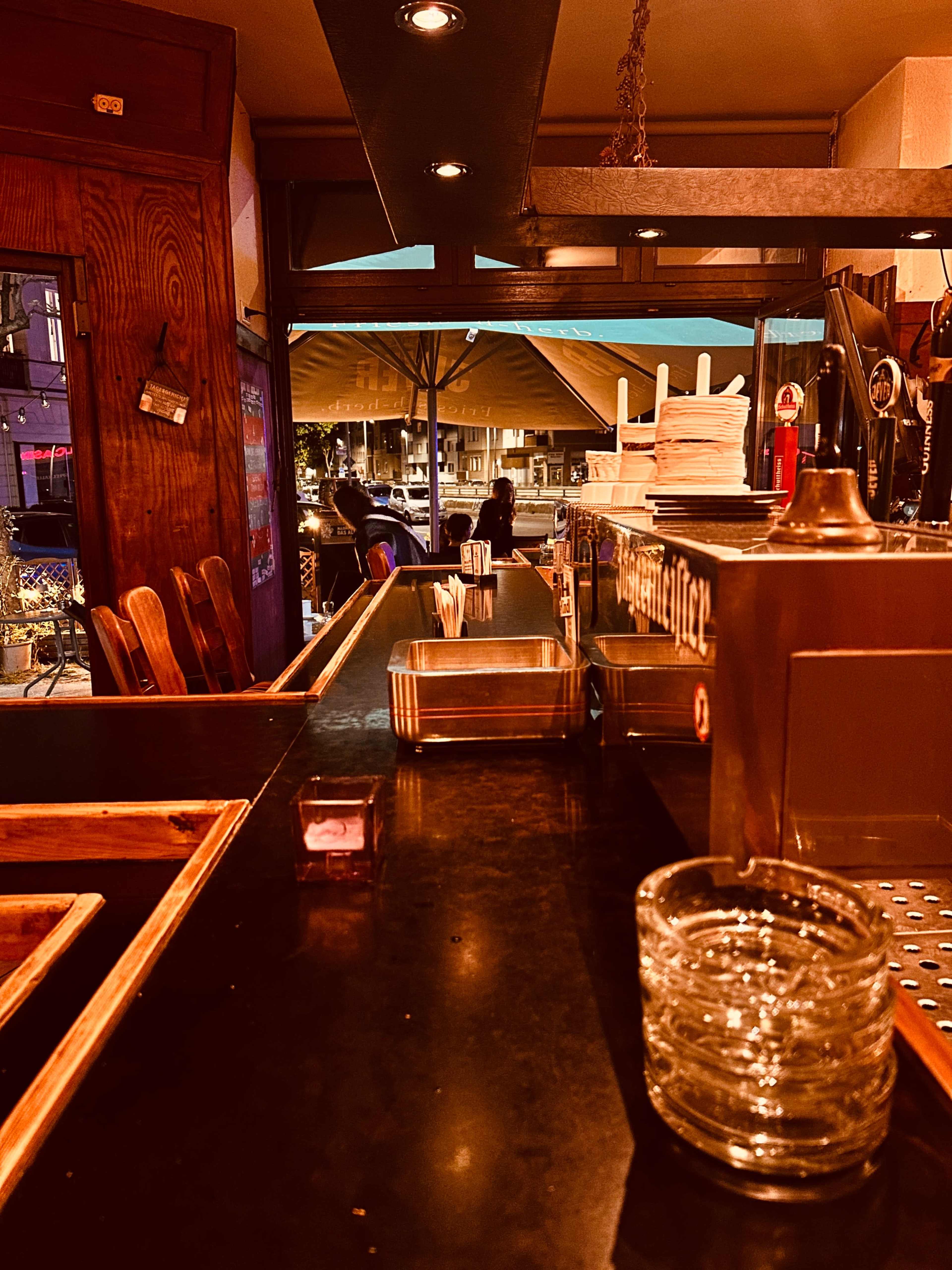The image shows a dimly lit bar with a polished counter, glassware, and a view of patrons sitting outside under a canopy.