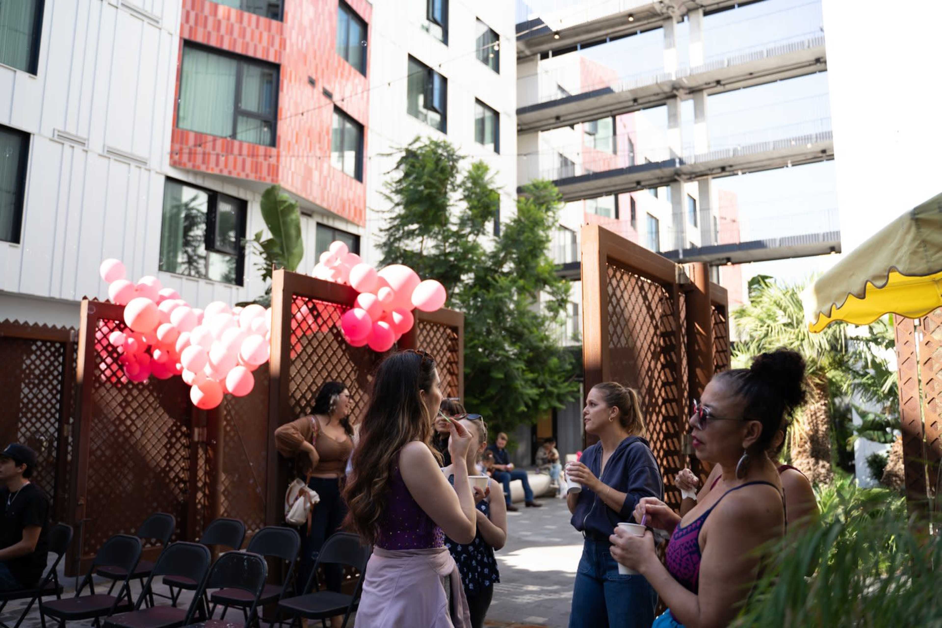 A group of people socializes in a courtyard decorated with pink balloons, surrounded by modern buildings and greenery.