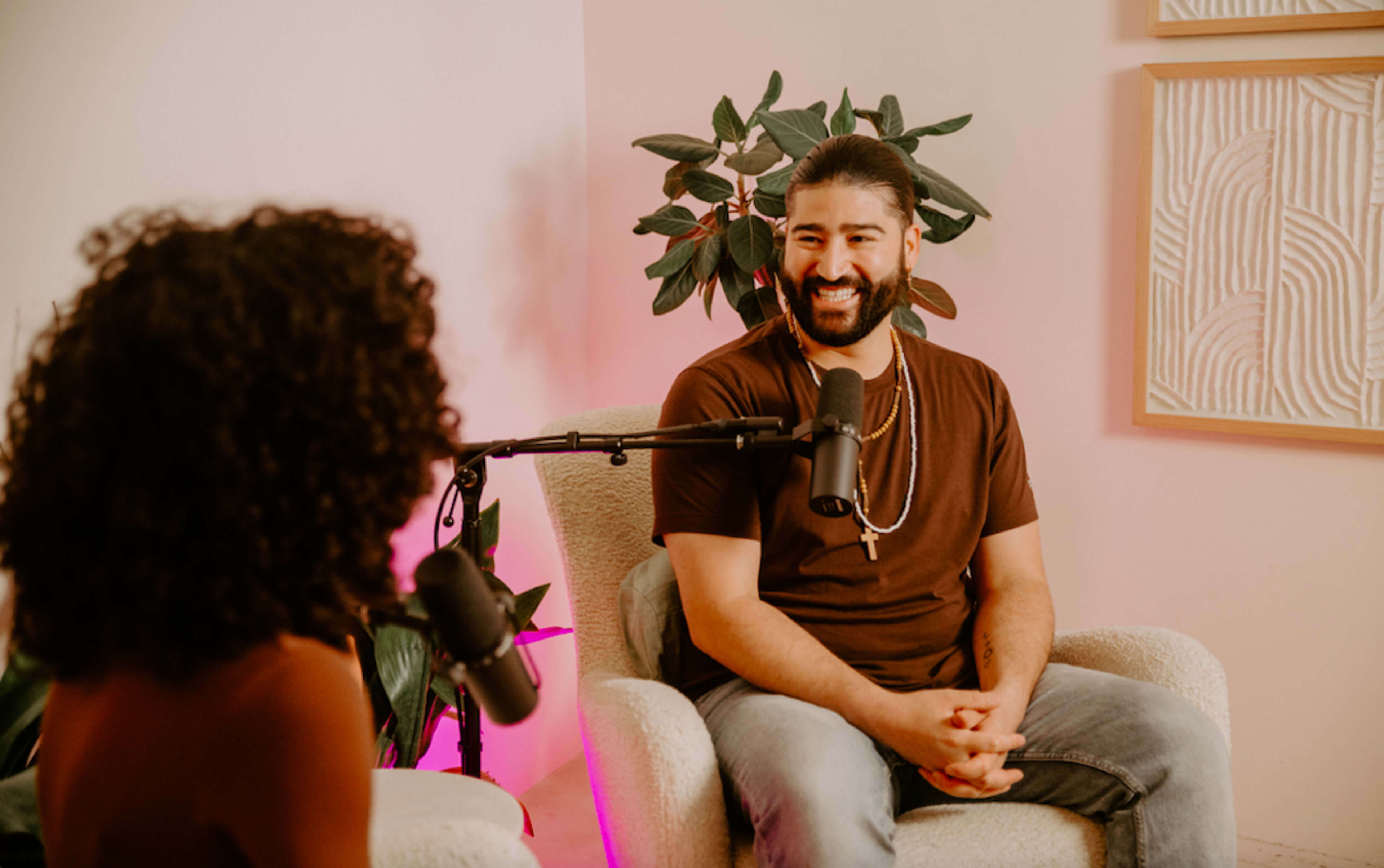 A man sits in a chair, smiling while speaking to a person partially visible across from him in a decorated studio setting.