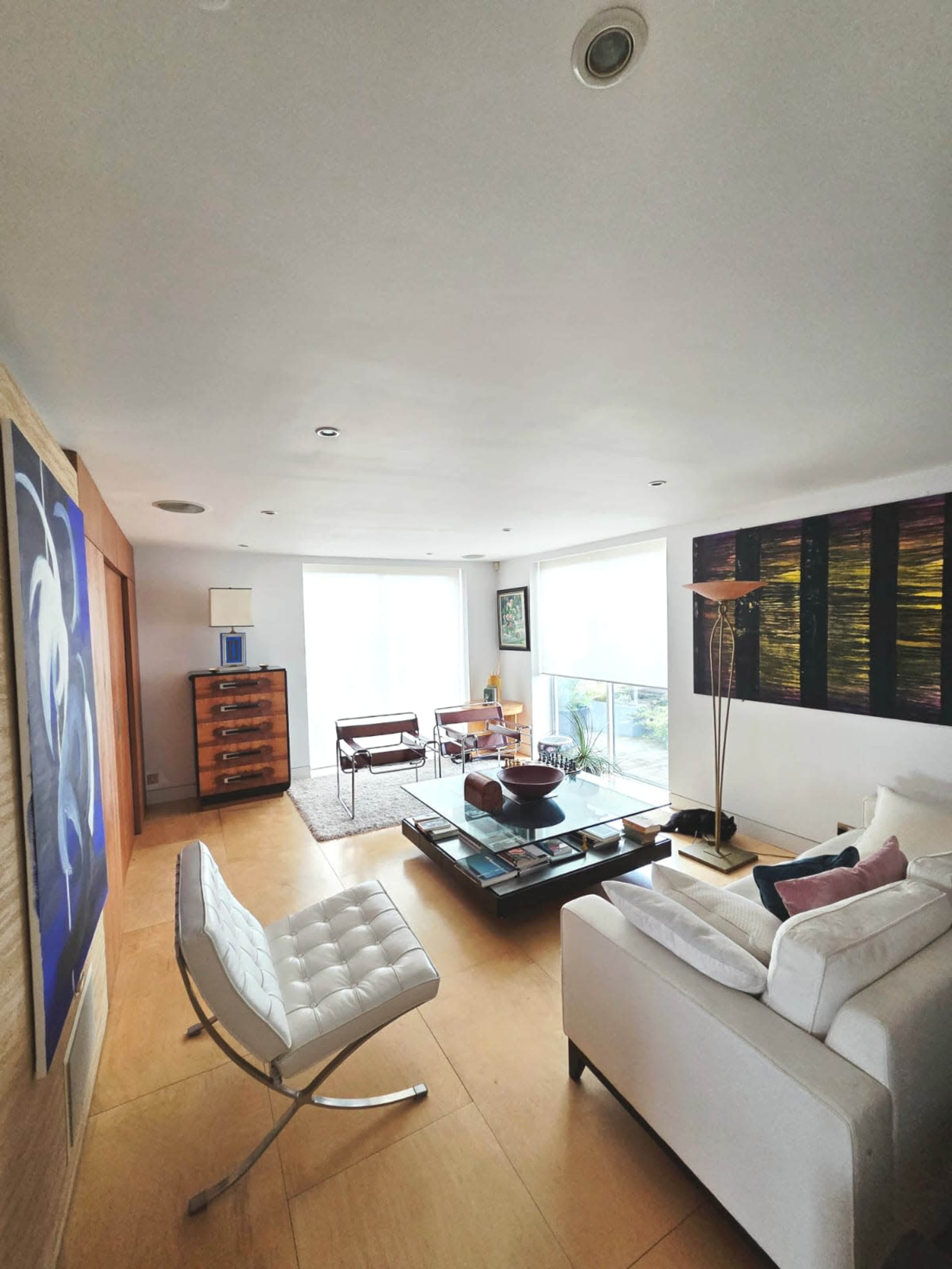 The image shows a modern living room with large windows, minimalist furniture, and an art piece on the wall.