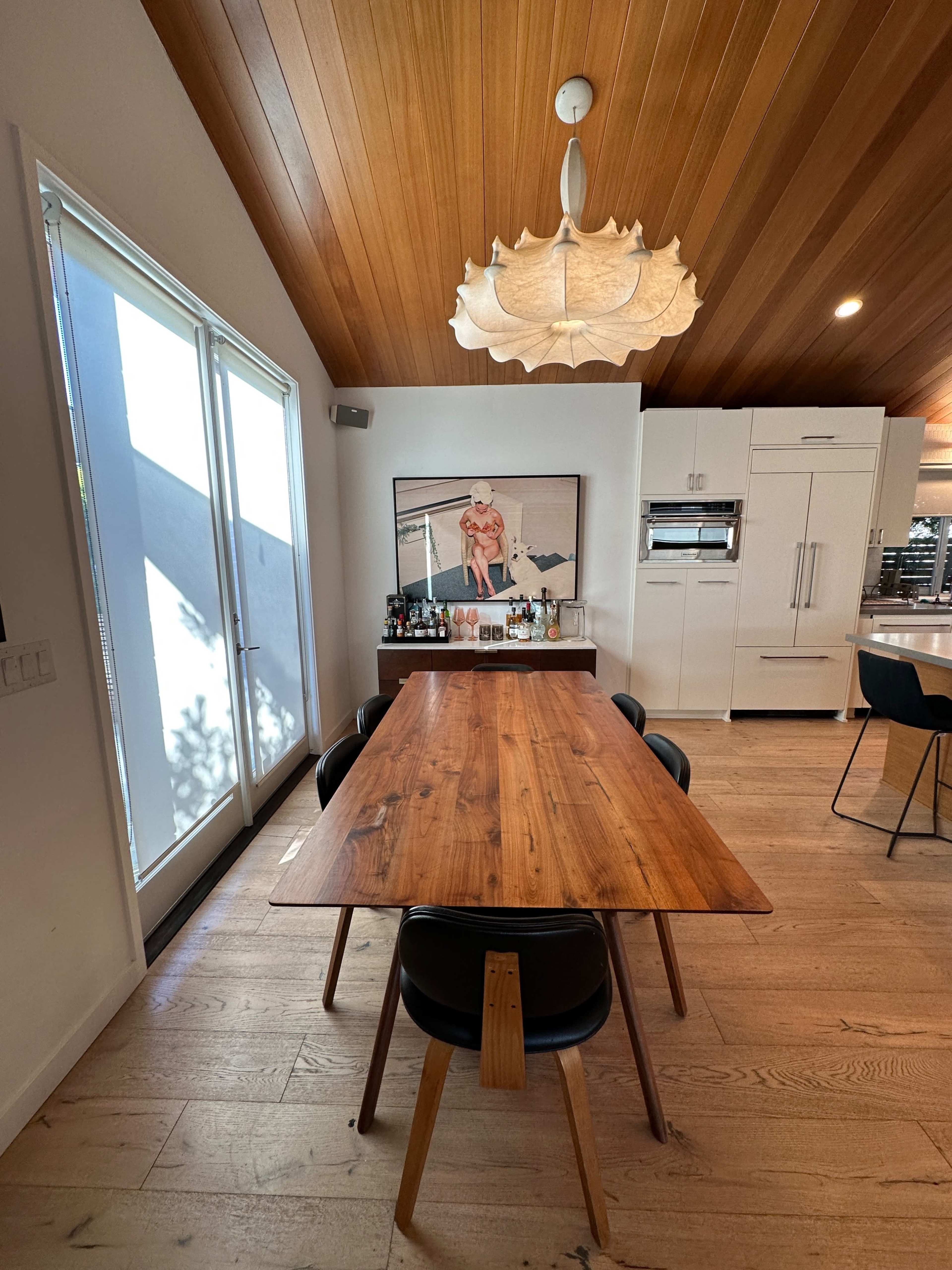 A wooden dining table with black chairs sits in a bright kitchen featuring large windows and a decorative light fixture above.