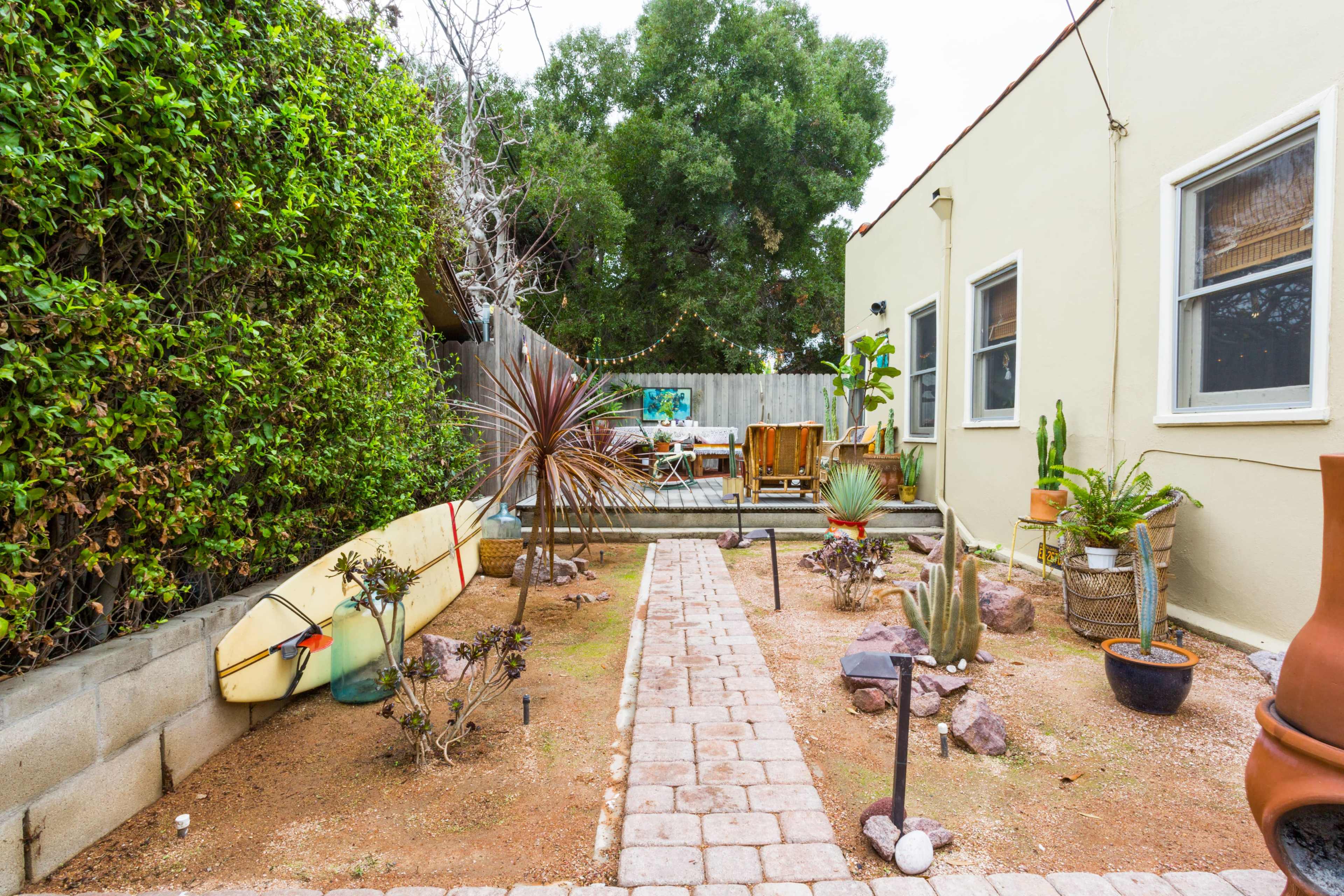 The image depicts a backyard garden with a stone pathway leading through various plants, including cacti, a surfboard, and outdoor furniture.