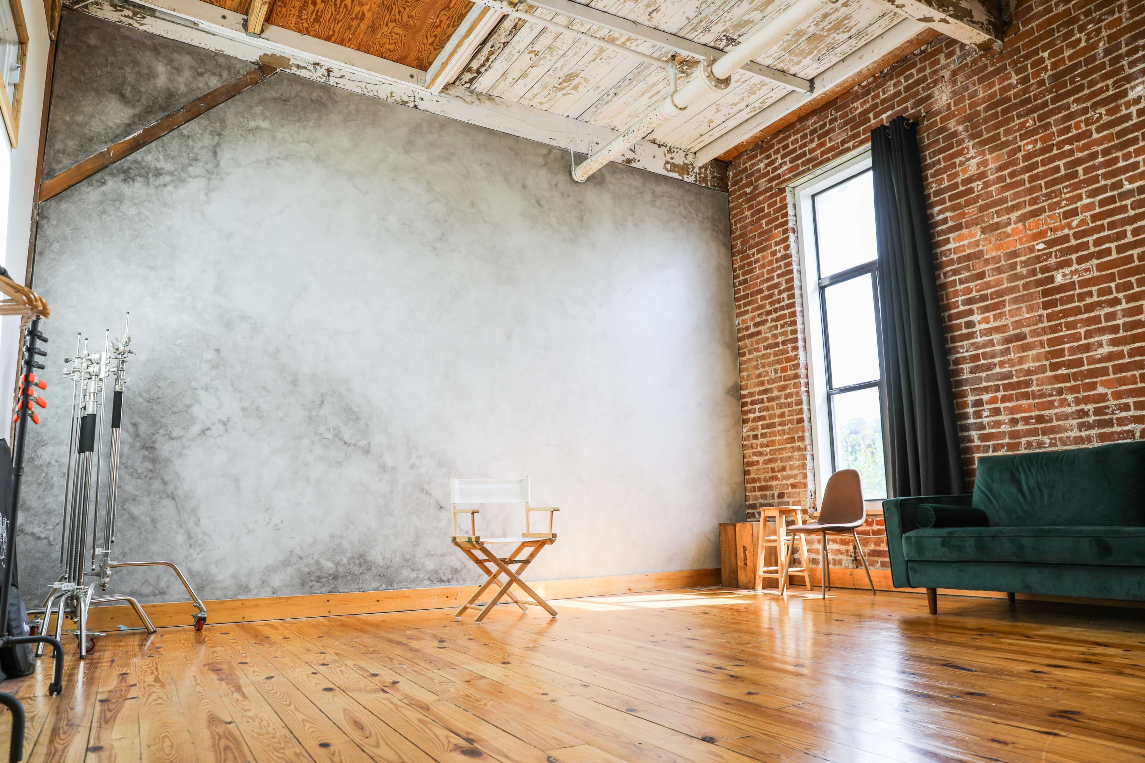 A spacious room with a wooden floor, exposed brick walls, a gray accent wall, a green sofa, a director's chair, and large windows that allow natural light to enter.