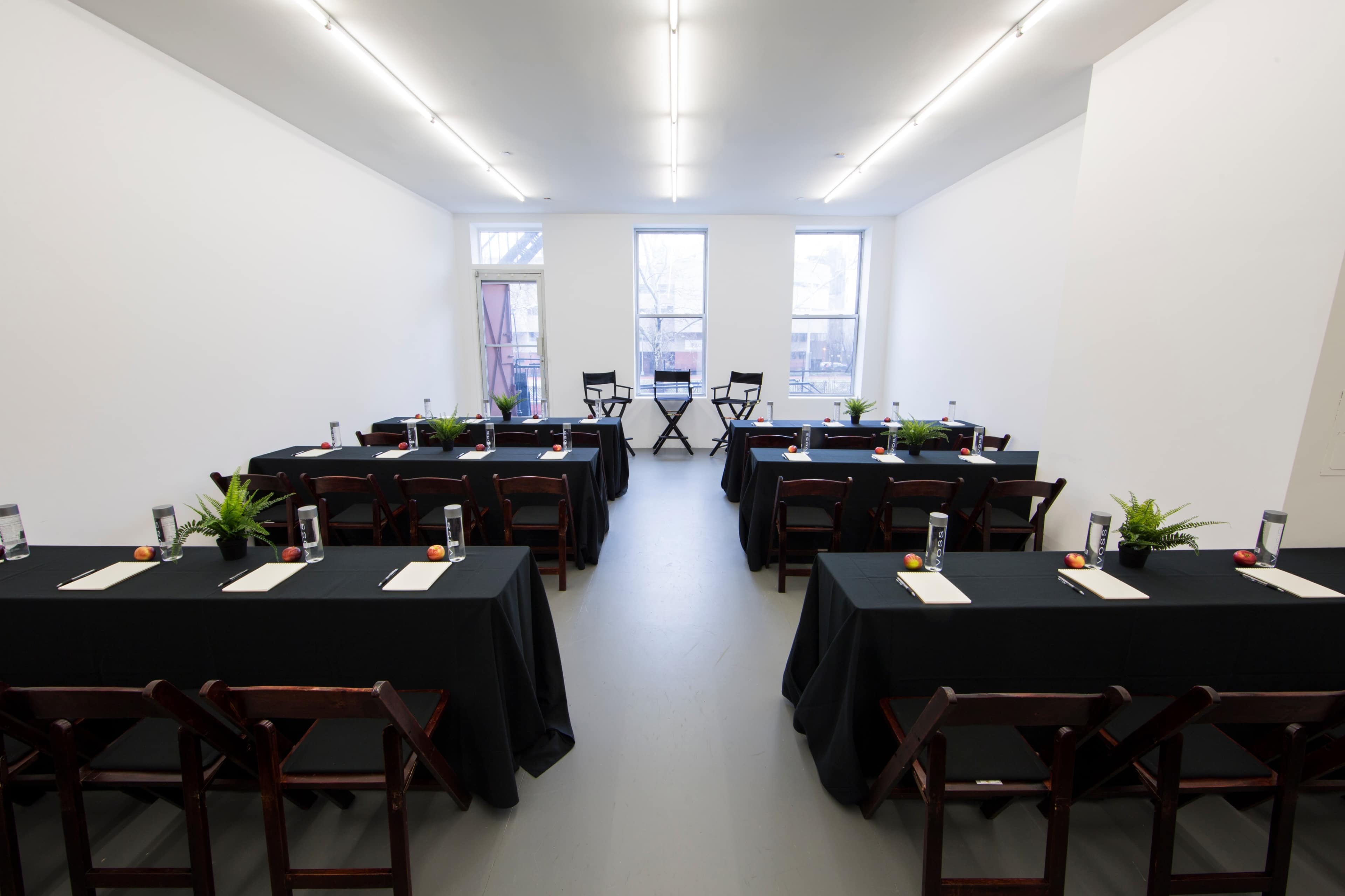 A spacious, empty room features two rows of tables with black tablecloths, each set with water bottles and writing materials, flanked by two chairs at the front.