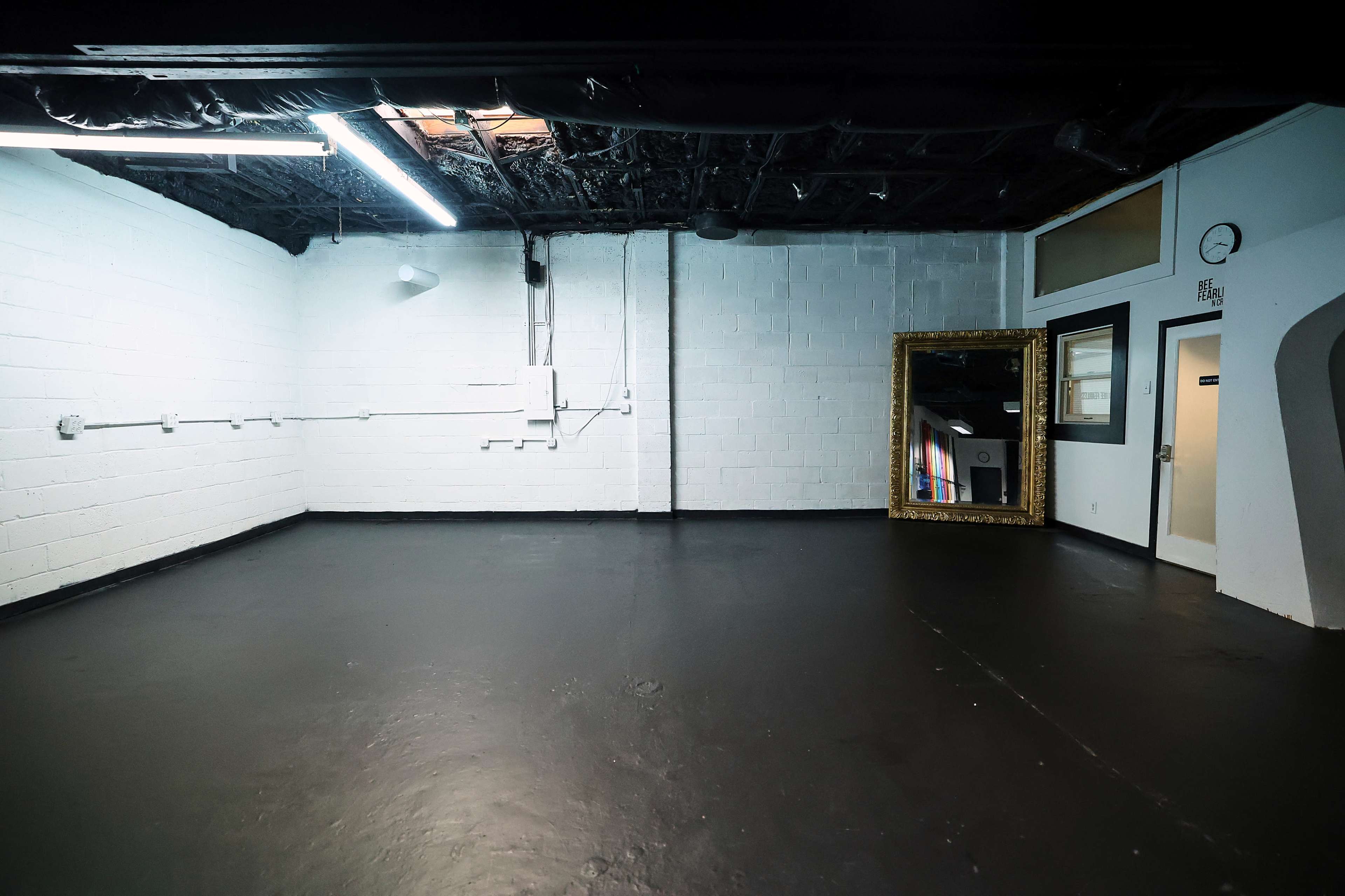 The image shows a sparsely furnished black-painted room with bare white walls, featuring a large ornate mirror against one wall and a clock near the ceiling.