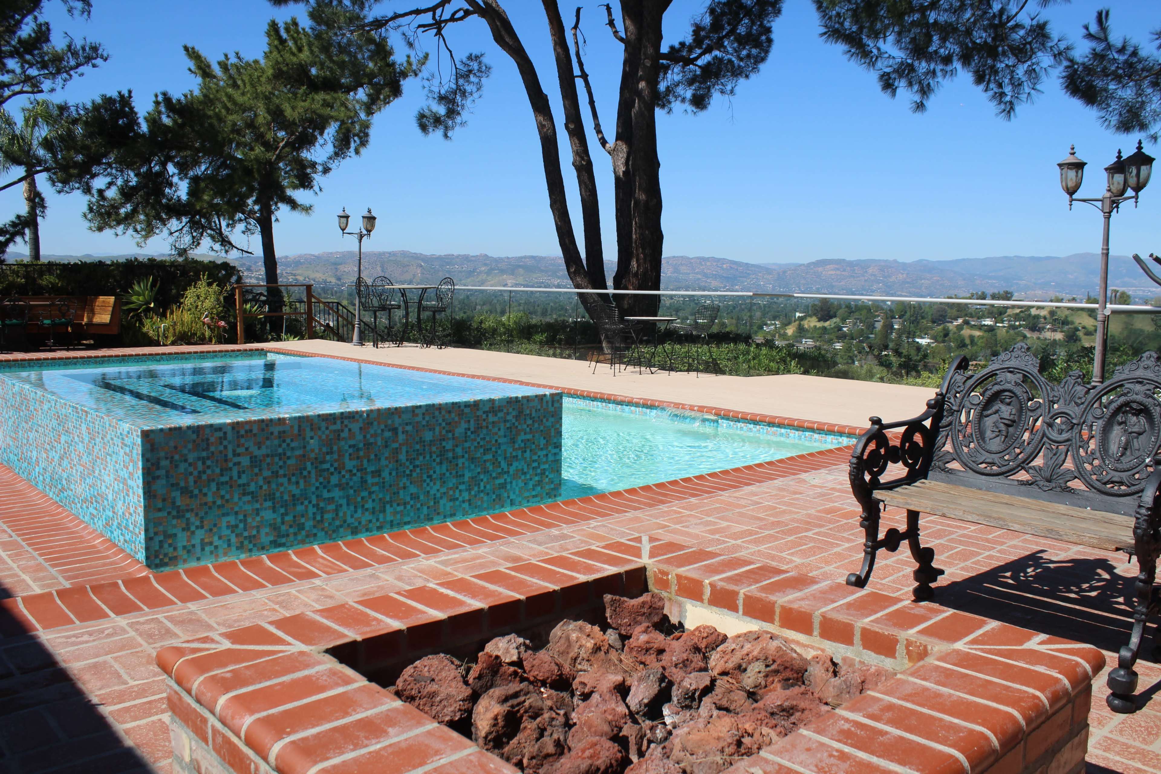 The image shows a pool area featuring two pools, one with mosaic tiles and a surrounding brick patio, against a backdrop of trees and a scenic view.