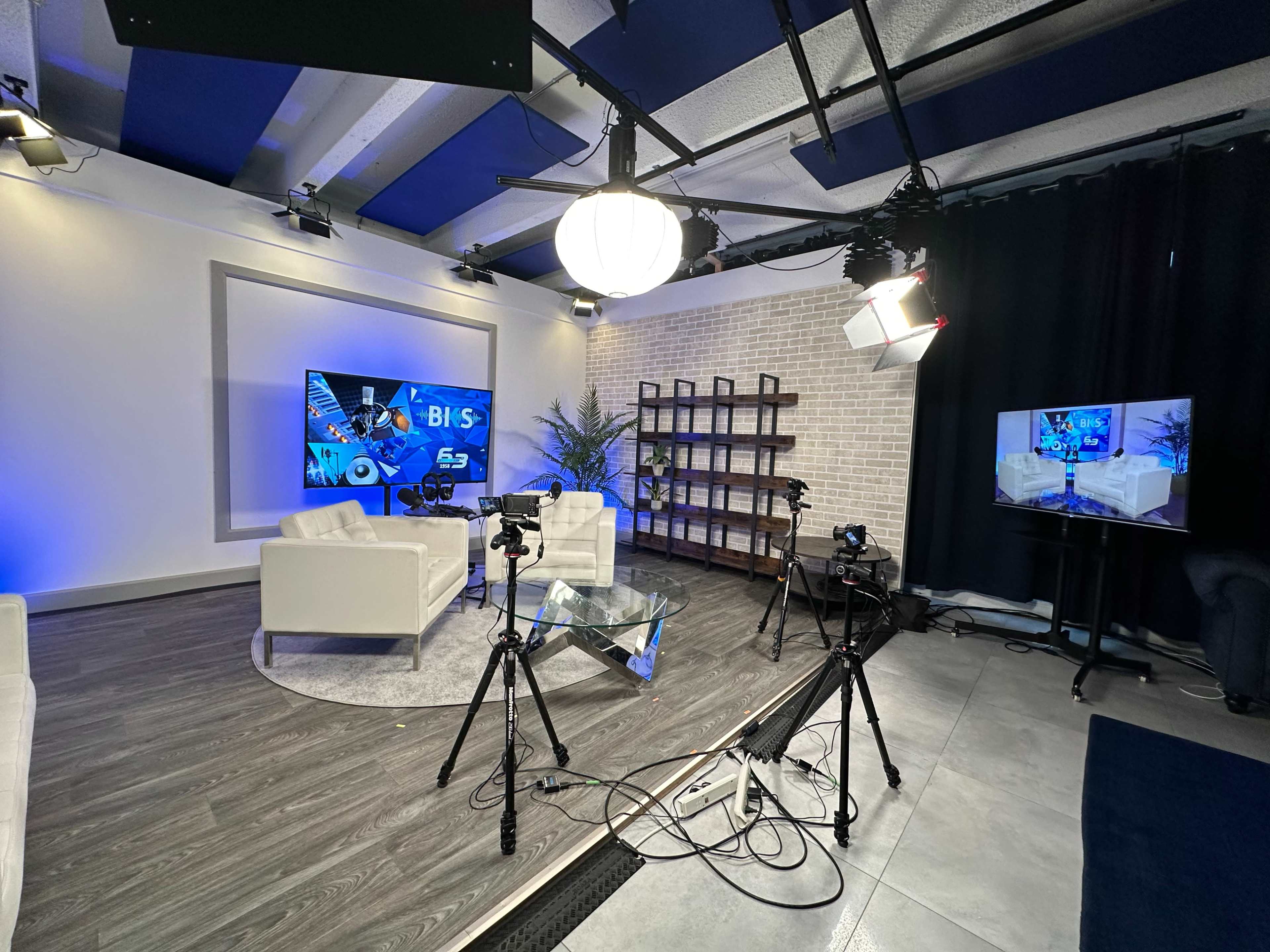 A modern studio setup with white sofas, a large screen displaying graphics, and various cameras positioned on tripods.