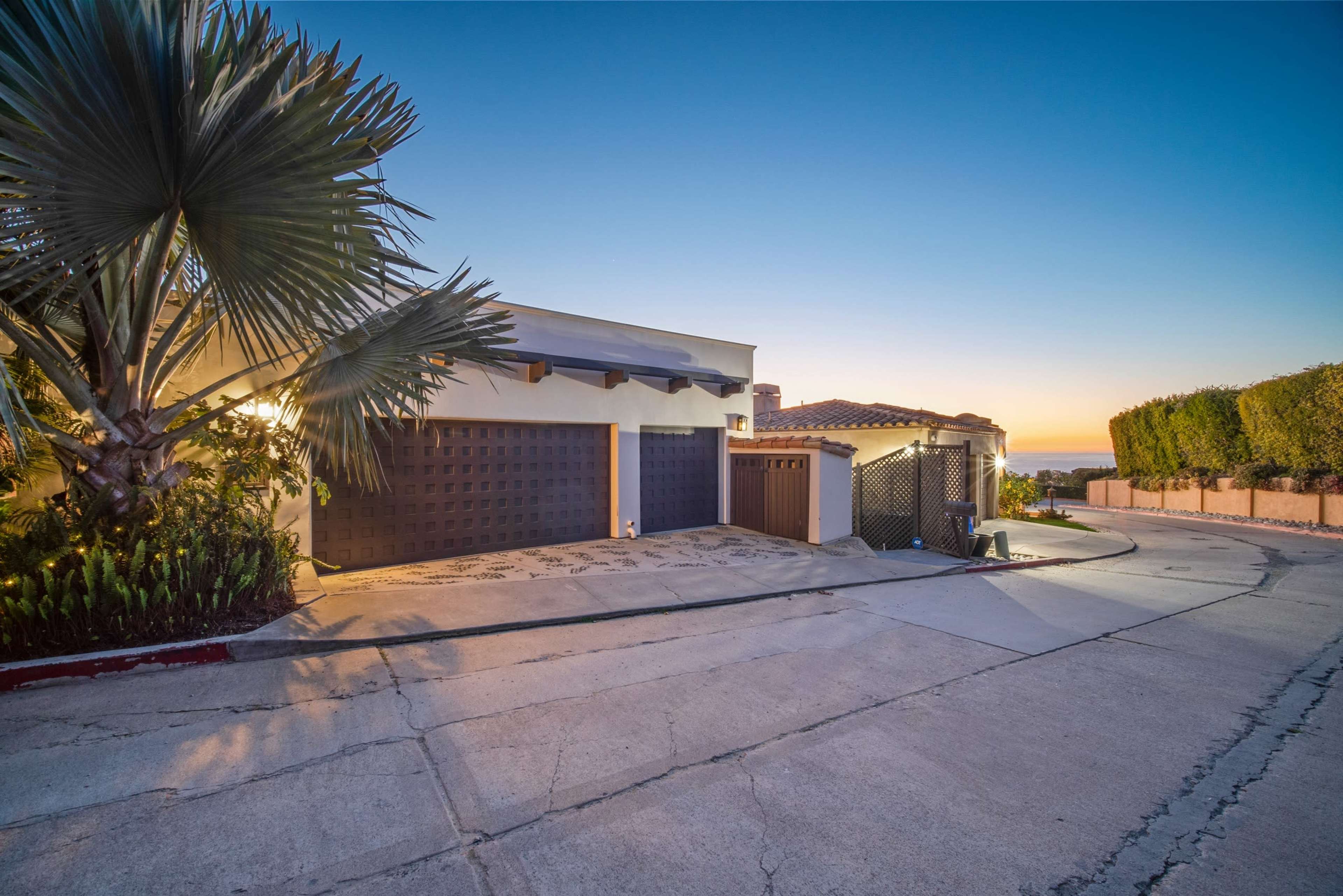 A modern house with a palm tree in the foreground, located on a curved driveway that slopes down toward the horizon at sunset.
