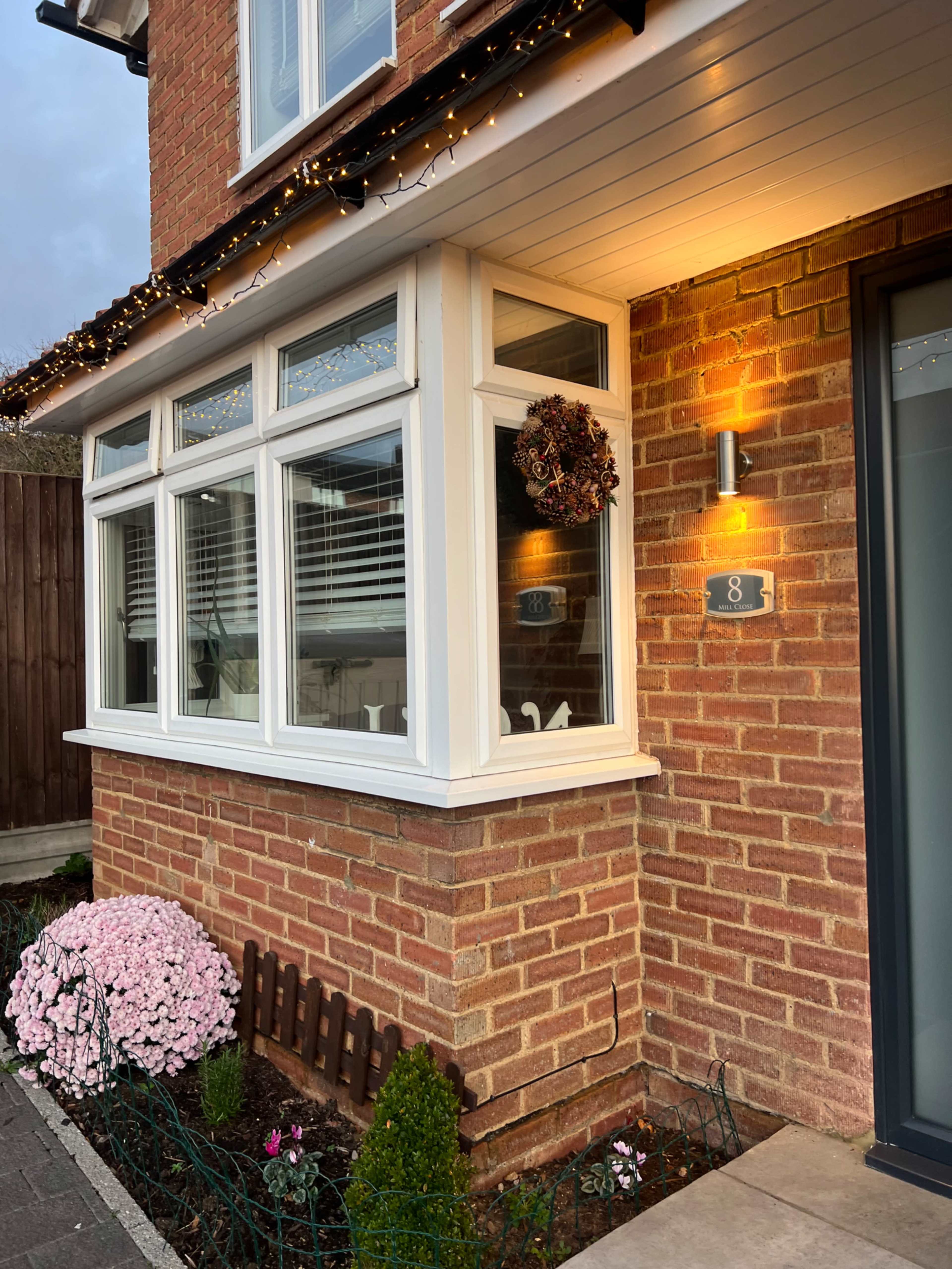 The image shows the corner of a brick house with white window frames, a decorative wreath on the window, and a well-maintained garden featuring pink flowers and a small wooden fence.