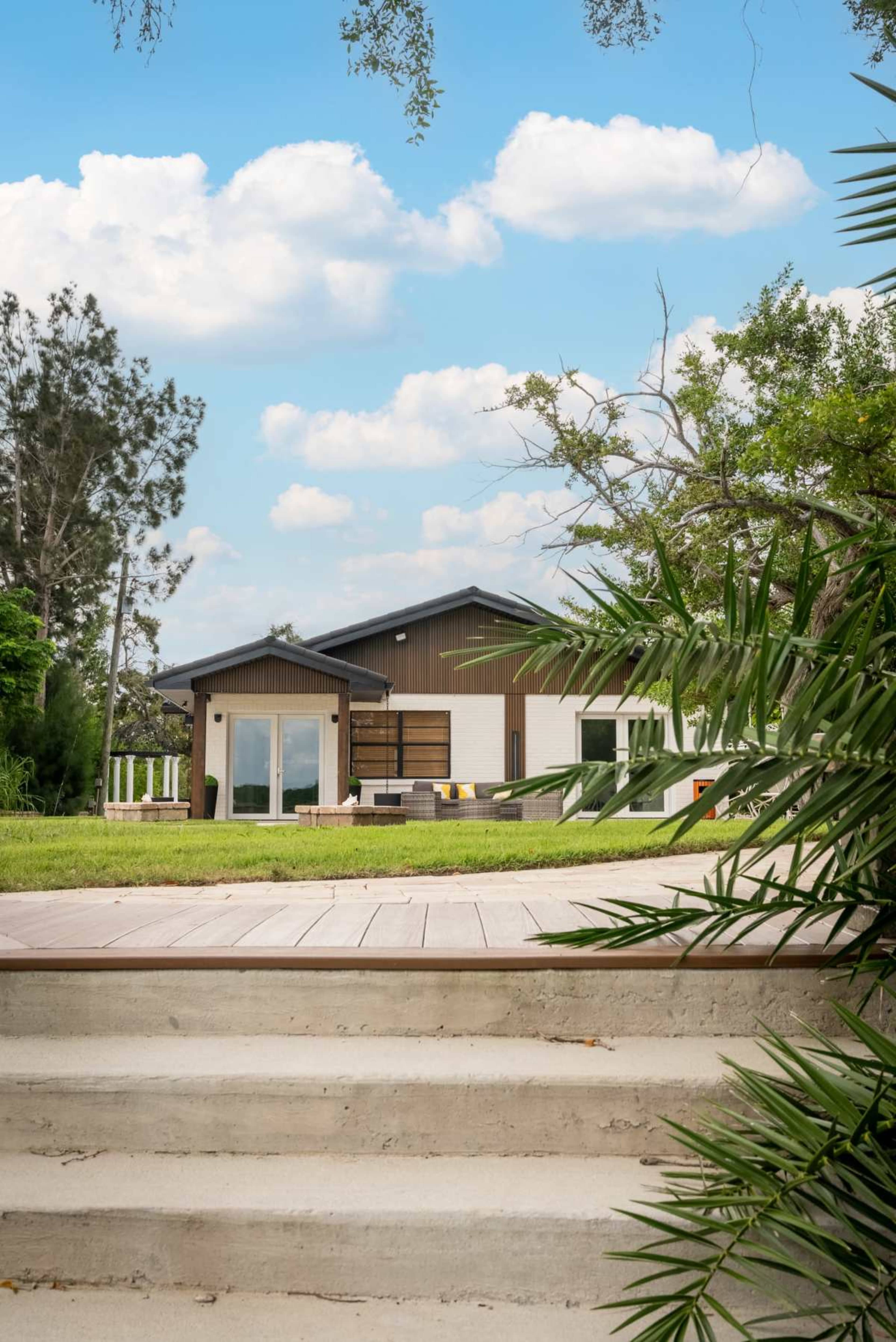 A modern house with a large yard and patio is surrounded by greenery and blue skies.