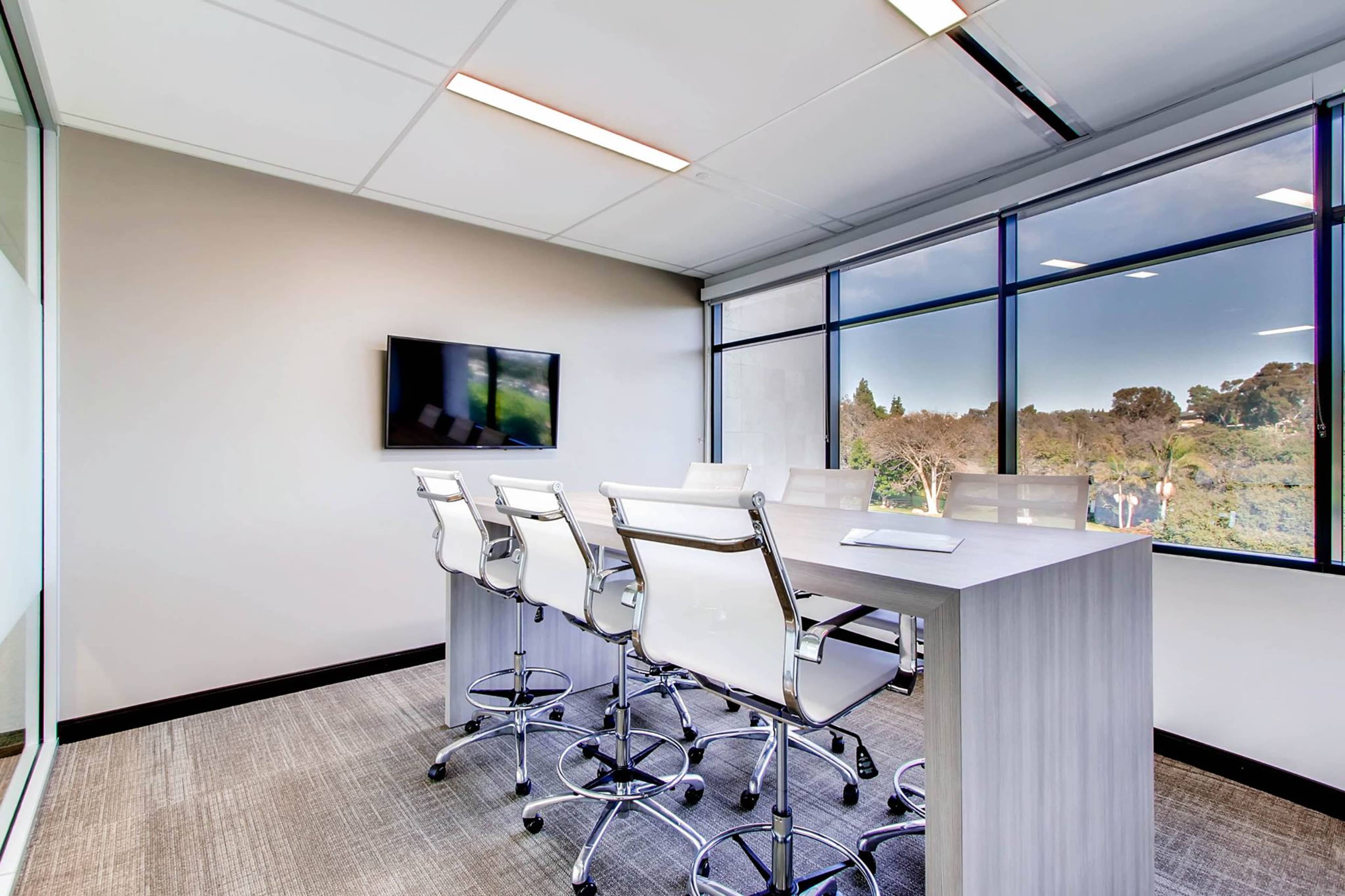 A modern conference room features a long table with high-backed chairs and a wall-mounted TV, surrounded by large windows offering a view of greenery.