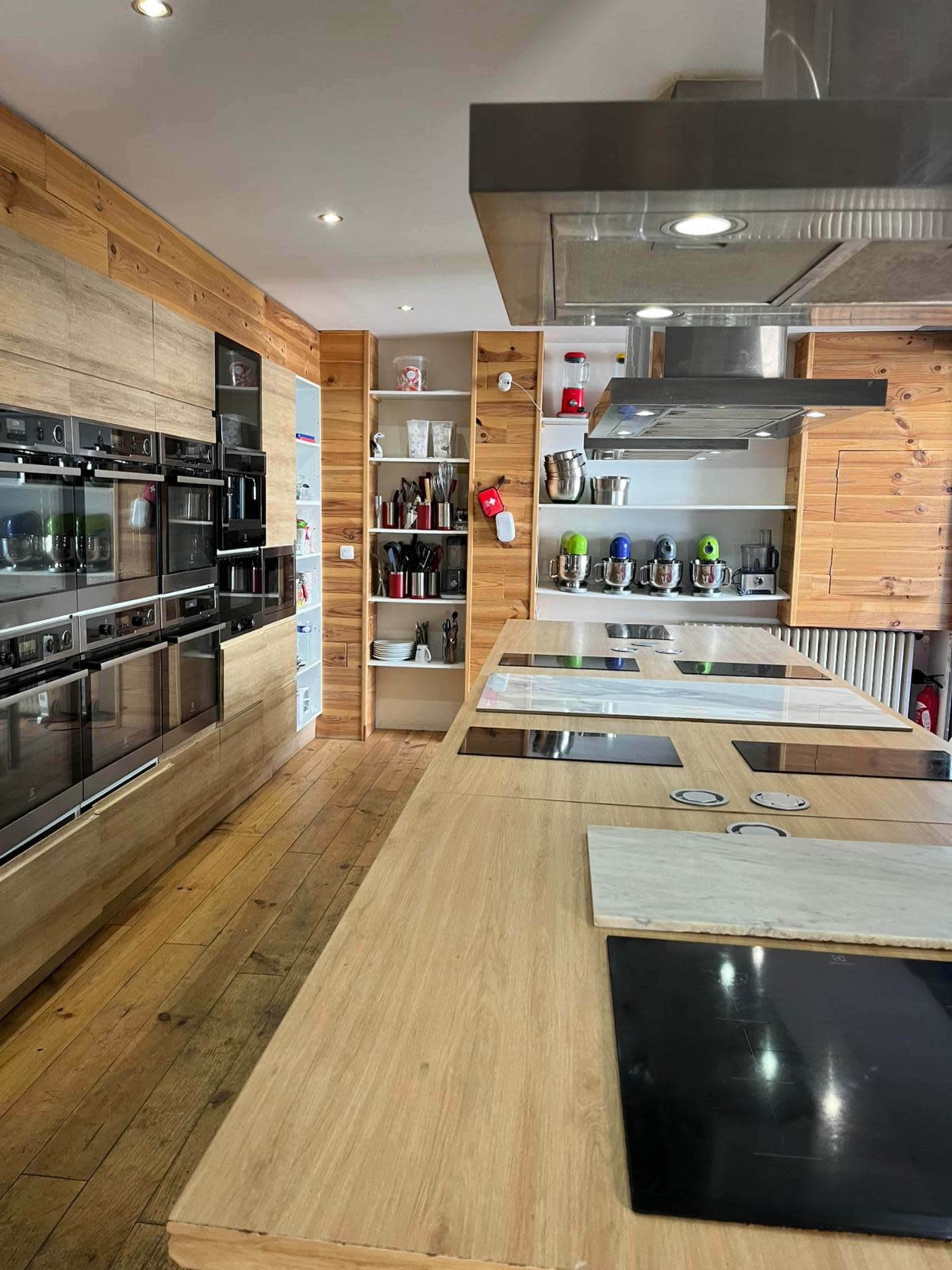 The image shows a modern kitchen with wooden cabinetry, multiple built-in ovens, and a large island featuring cooking surfaces.