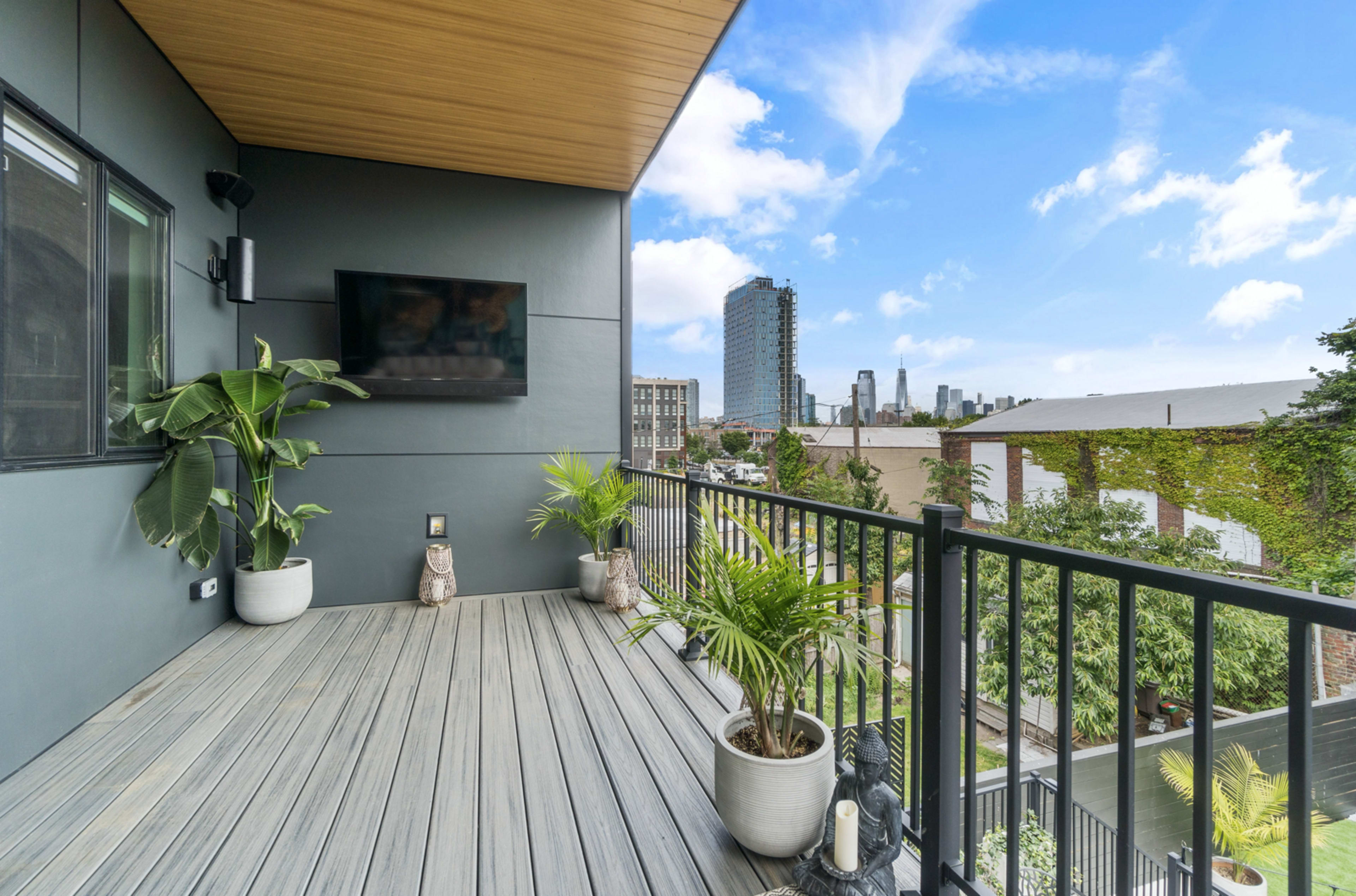 A modern balcony with wooden flooring, potted plants, and a city skyline visible in the background.