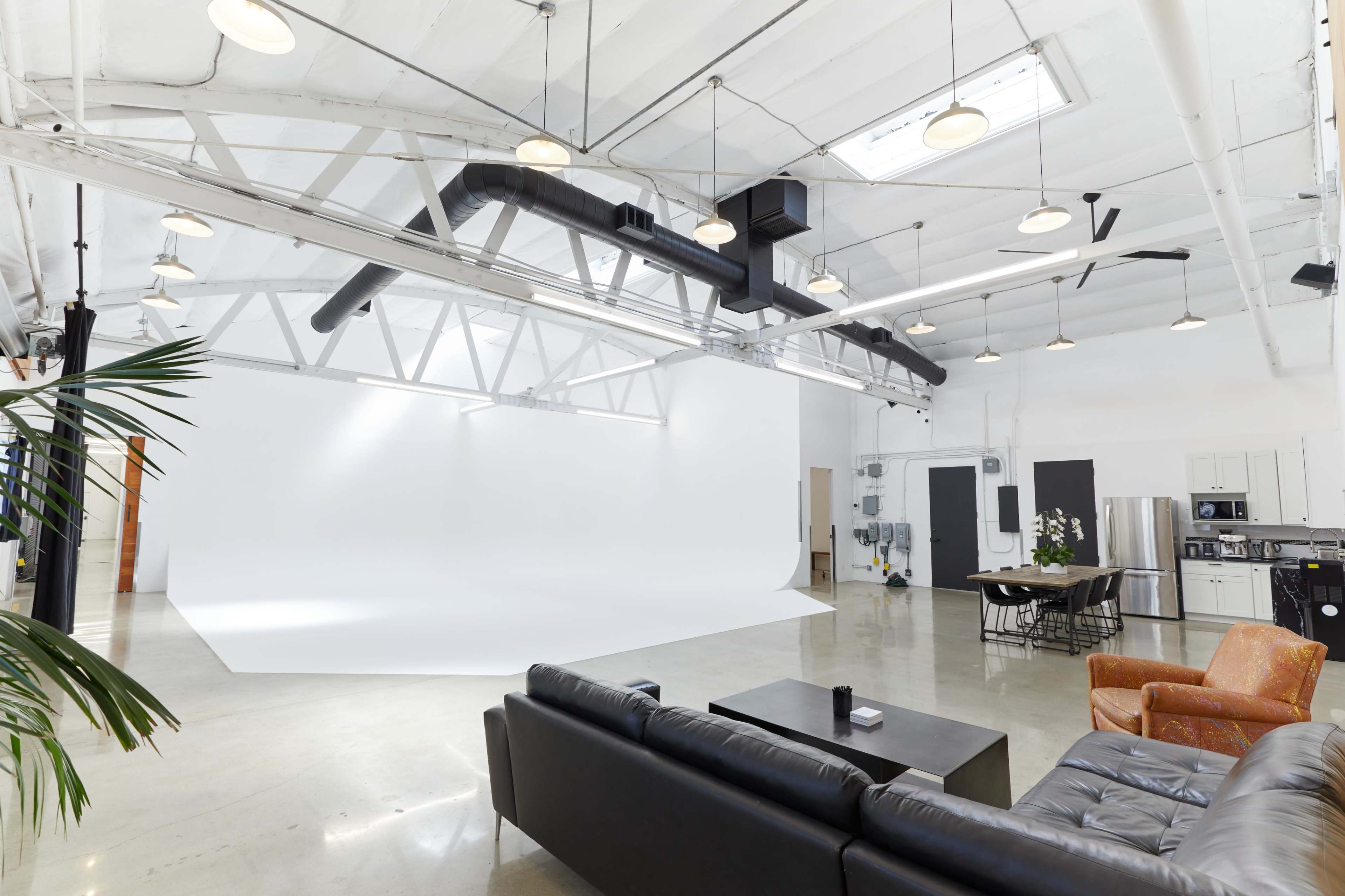 The image shows a spacious, bright studio with a white backdrop, a black couch, an orange armchair, and a dining table with chairs.