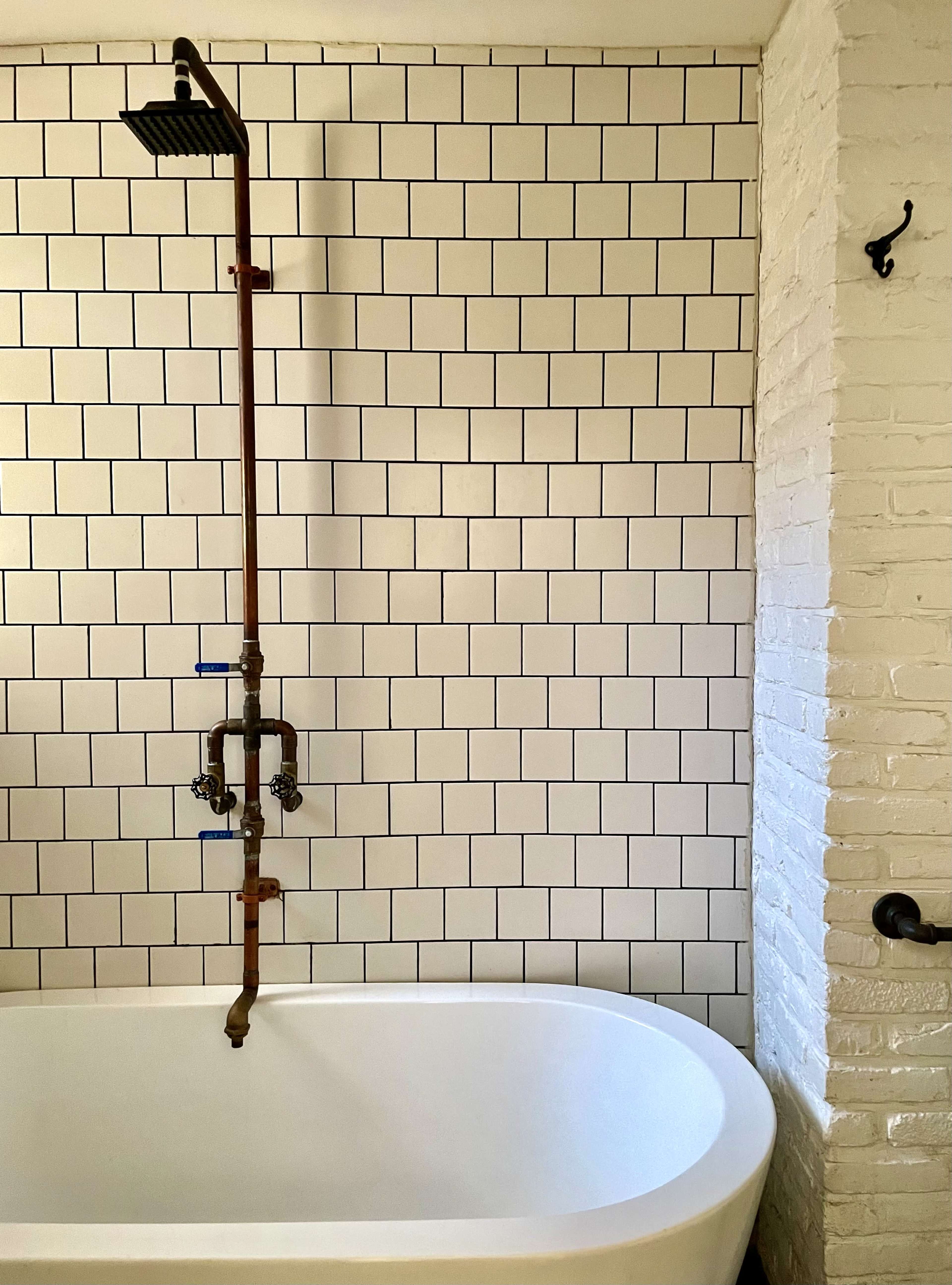 The image shows a modern bathroom featuring a white bathtub beneath a copper shower fixture attached to a tiled wall with rectangular white tiles.