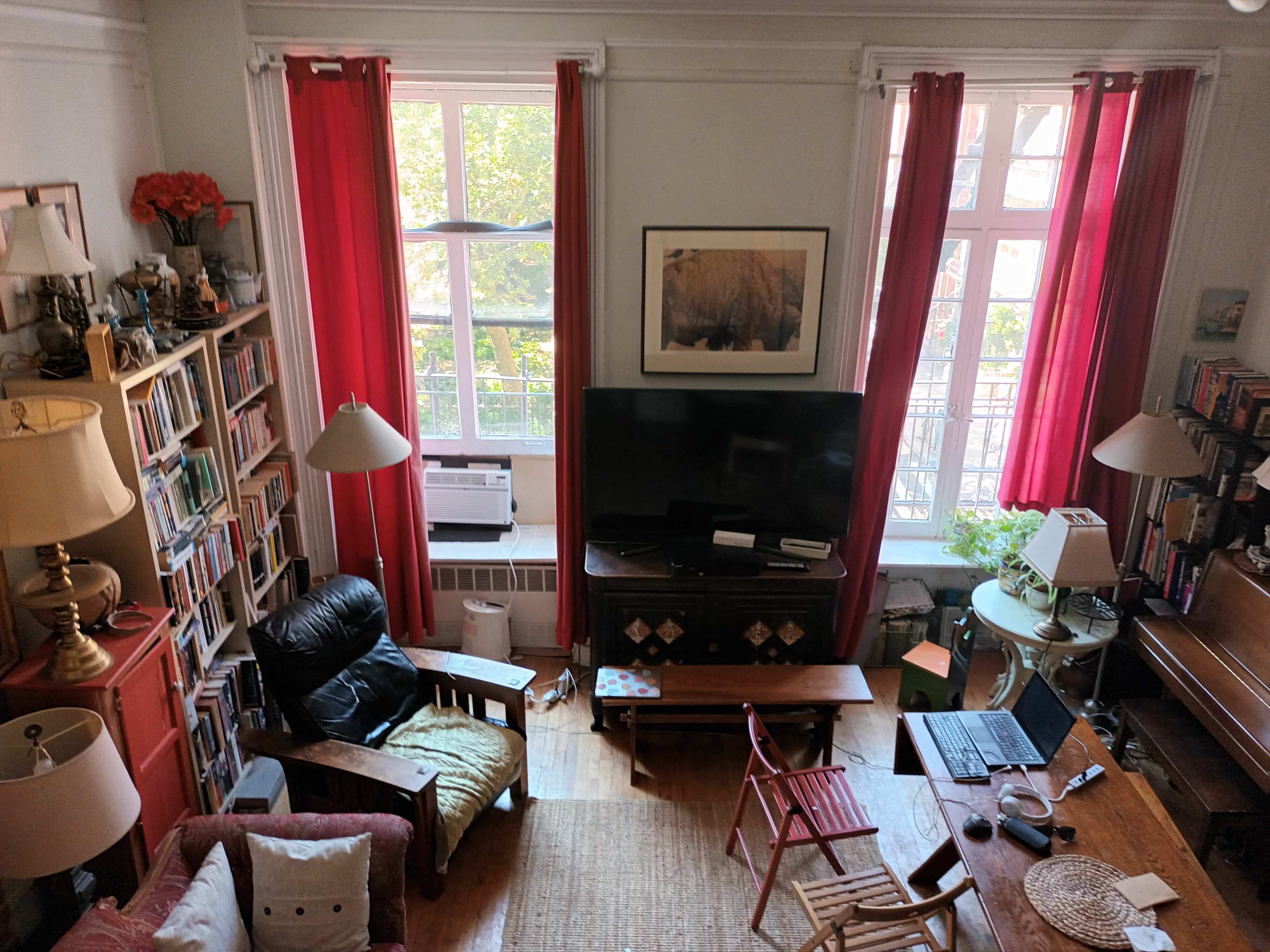 A cozy living room with two large windows, a television stand, a bookshelf filled with books, and various furniture items including a black chair and a wooden table.