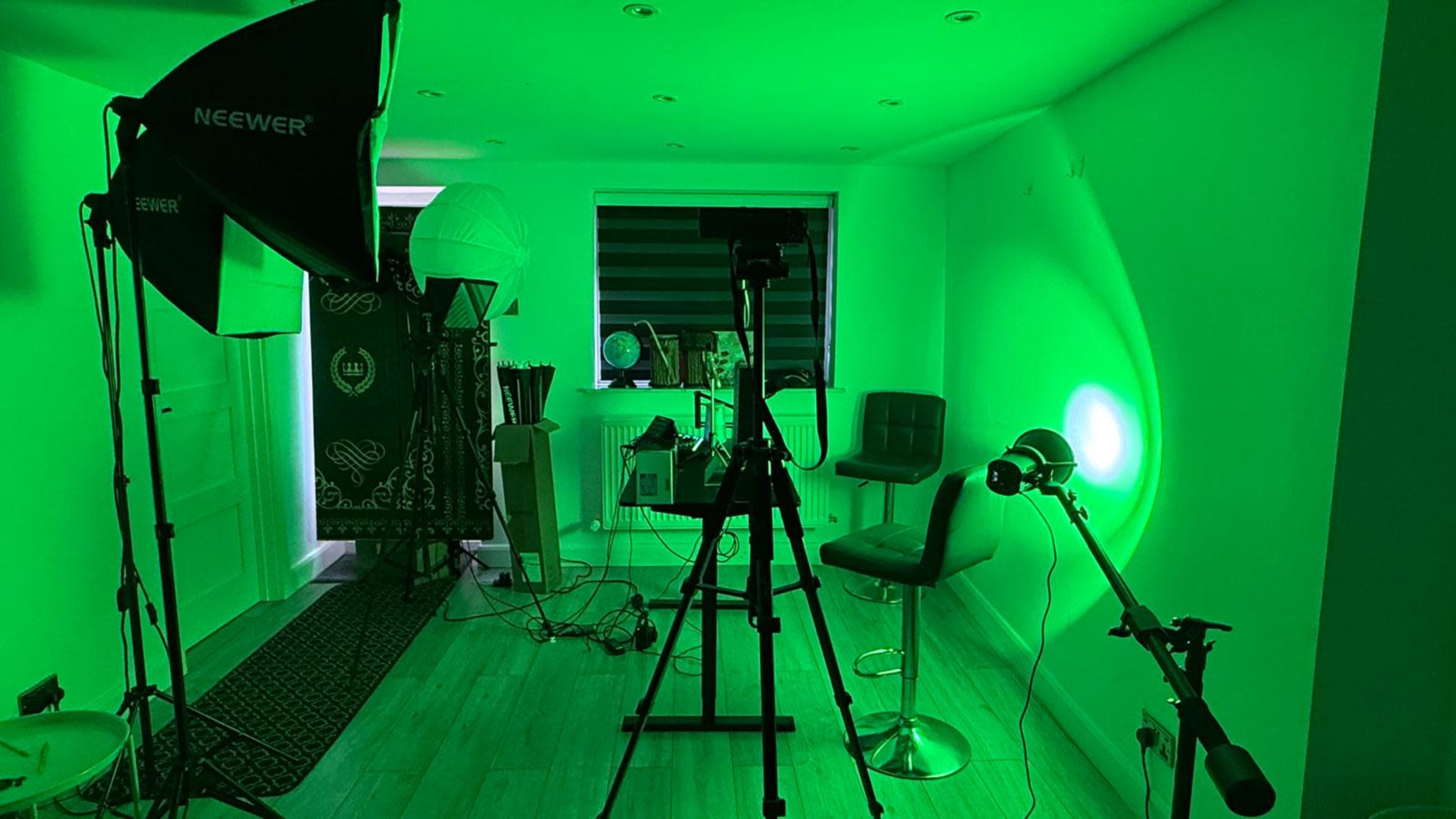 The image shows a photography or videography studio with green lighting, featuring a camera on a tripod, a backdrop, and various studio equipment arranged in the room.