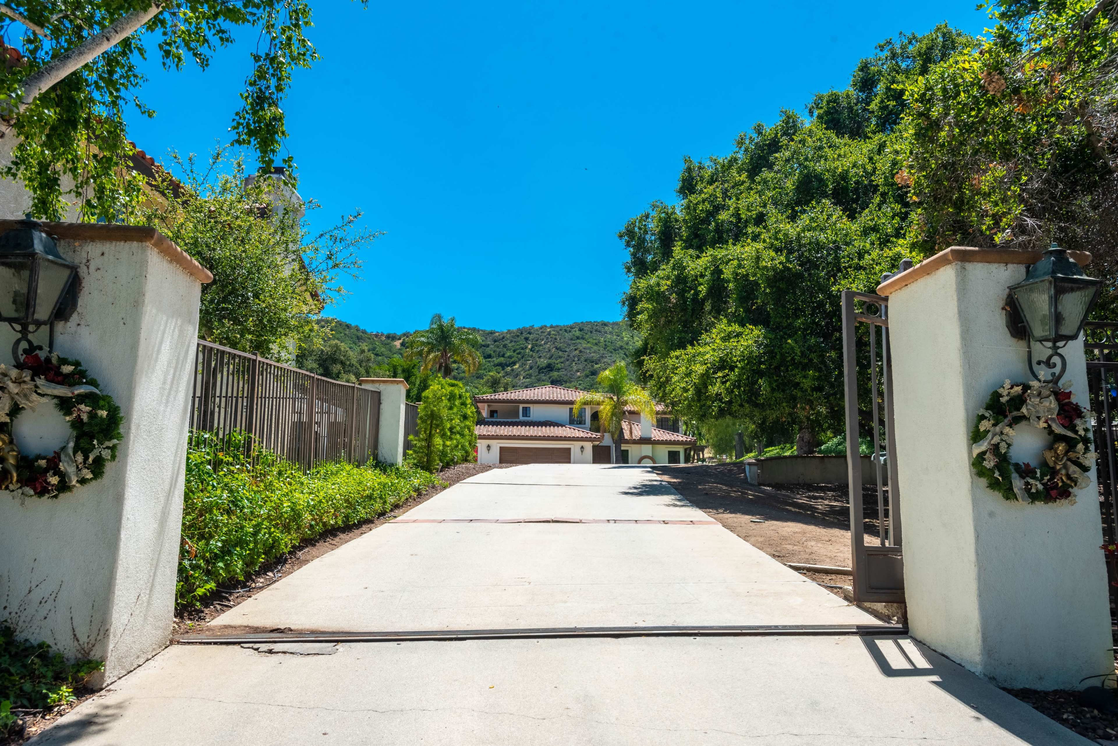 A long, paved driveway leads to a house set among trees and mountains, with decorative lanterns on either side of the entrance.