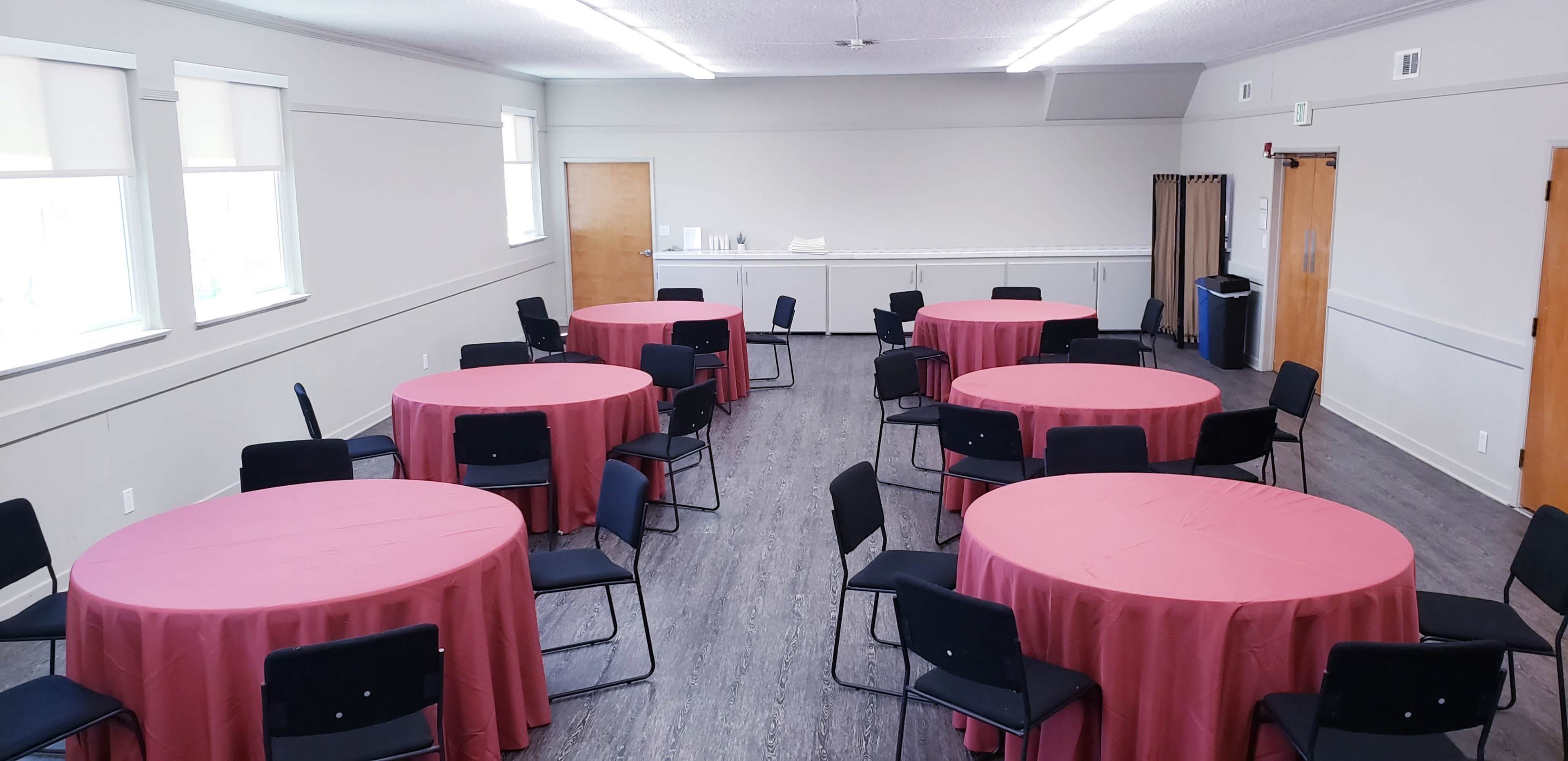 A room with light gray walls contains several round tables covered with pink tablecloths, surrounded by black chairs.