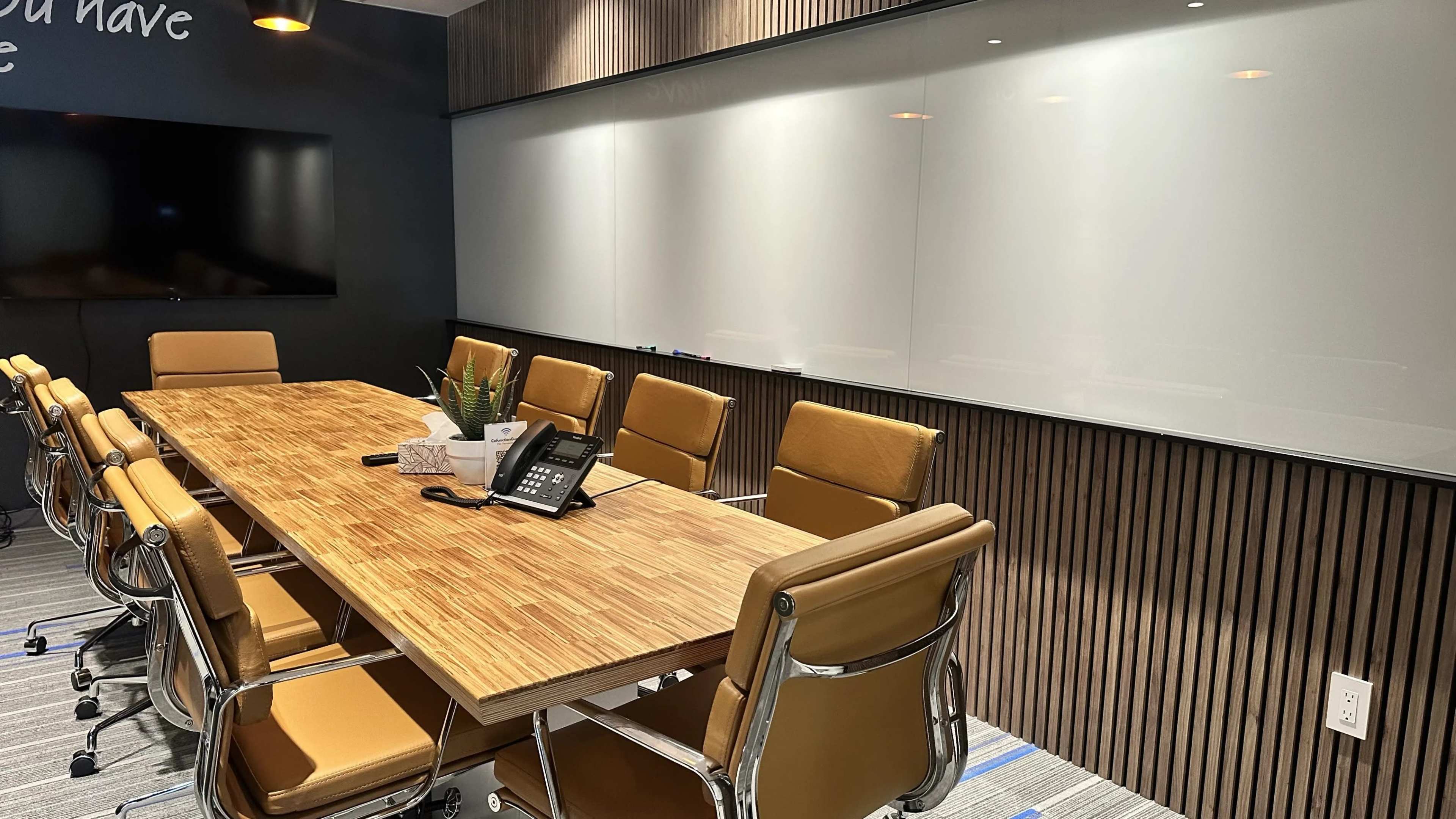 The image shows a modern conference room with a large wooden table, several leather chairs, a television screen on the wall, and a whiteboard.