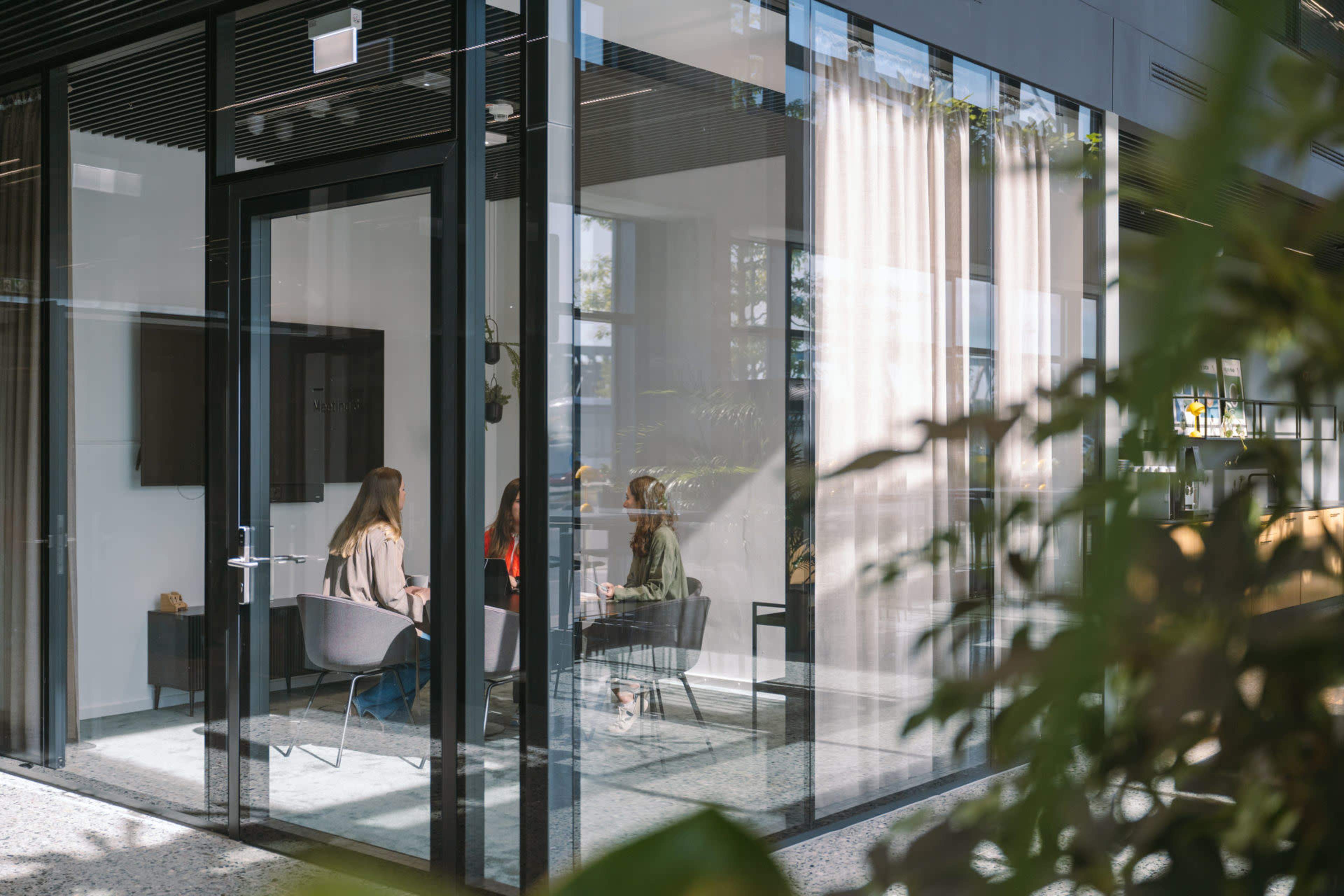 Three people are seated in a glass-walled meeting room with greenery visible outside.