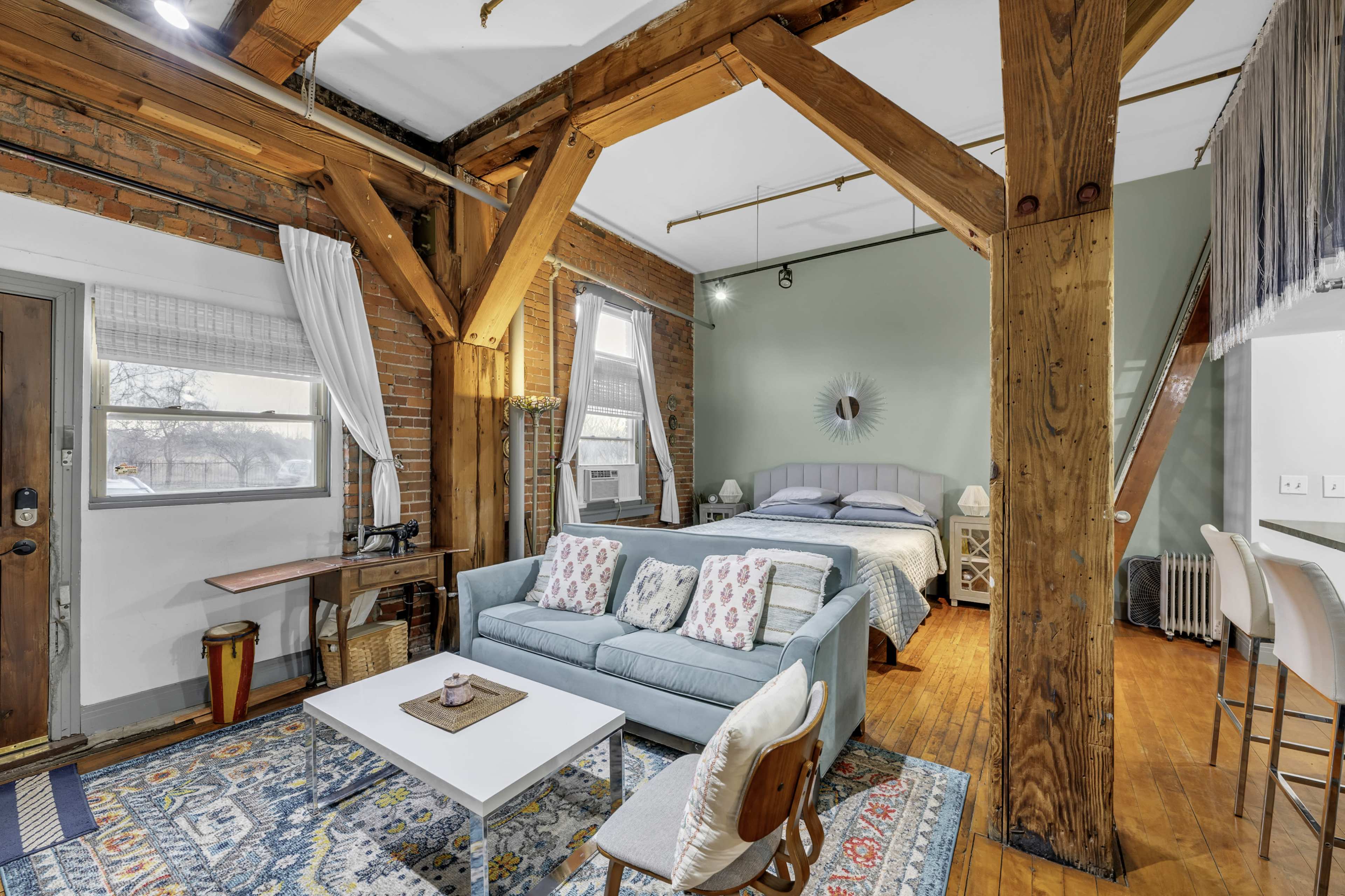 The image shows a cozy living space featuring exposed wooden beams, a light blue sofa, a small coffee table, and a bed in the background against a green wall.
