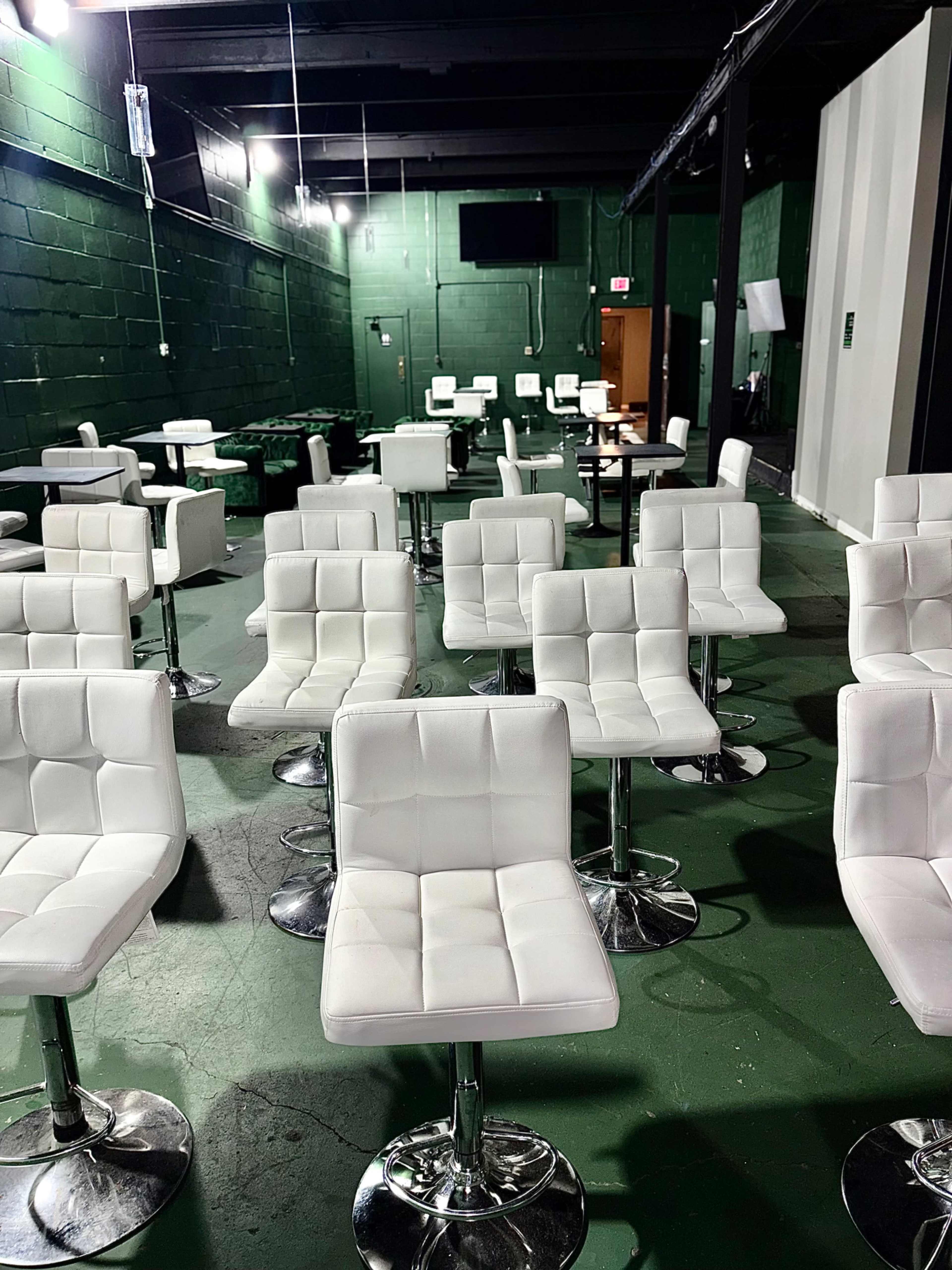 The image shows a room filled with white, modern swivel chairs arranged neatly on a green floor, with sparse lighting and a green-painted wall.