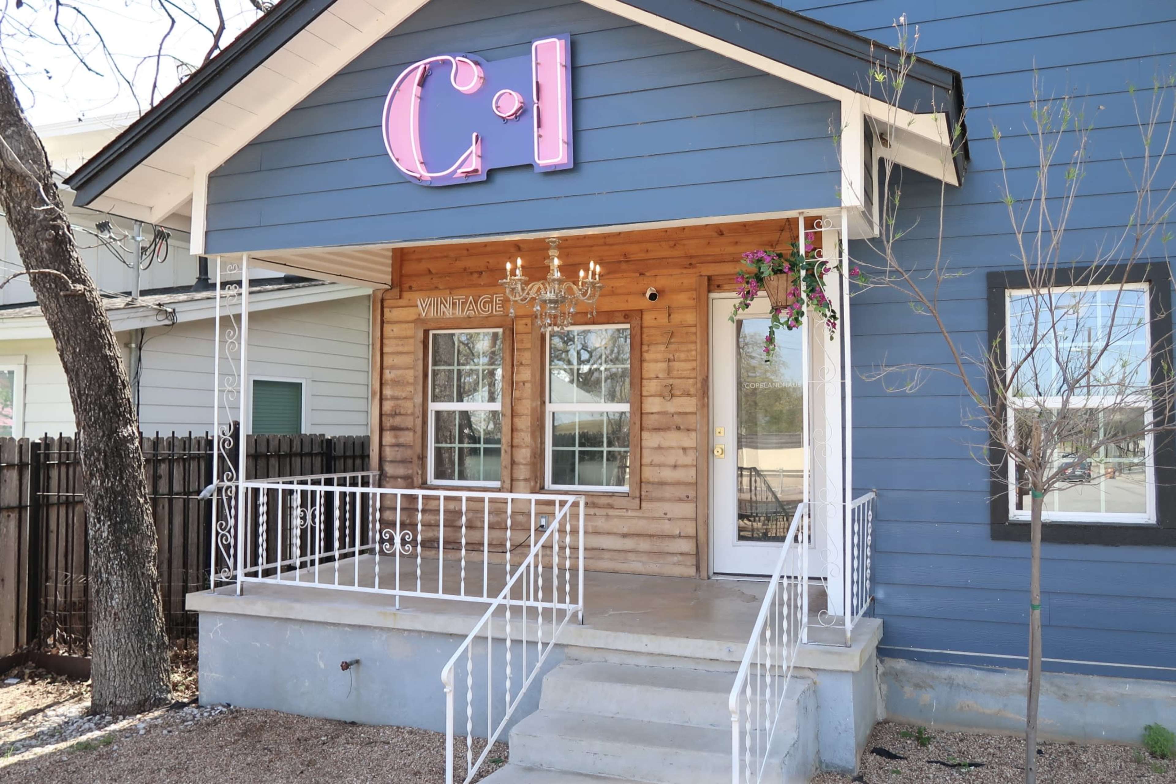 The image shows a blue house with a wooden section, featuring a sign that says "C1" in pink neon, and a front porch adorned with a chandelier and hanging plants.
