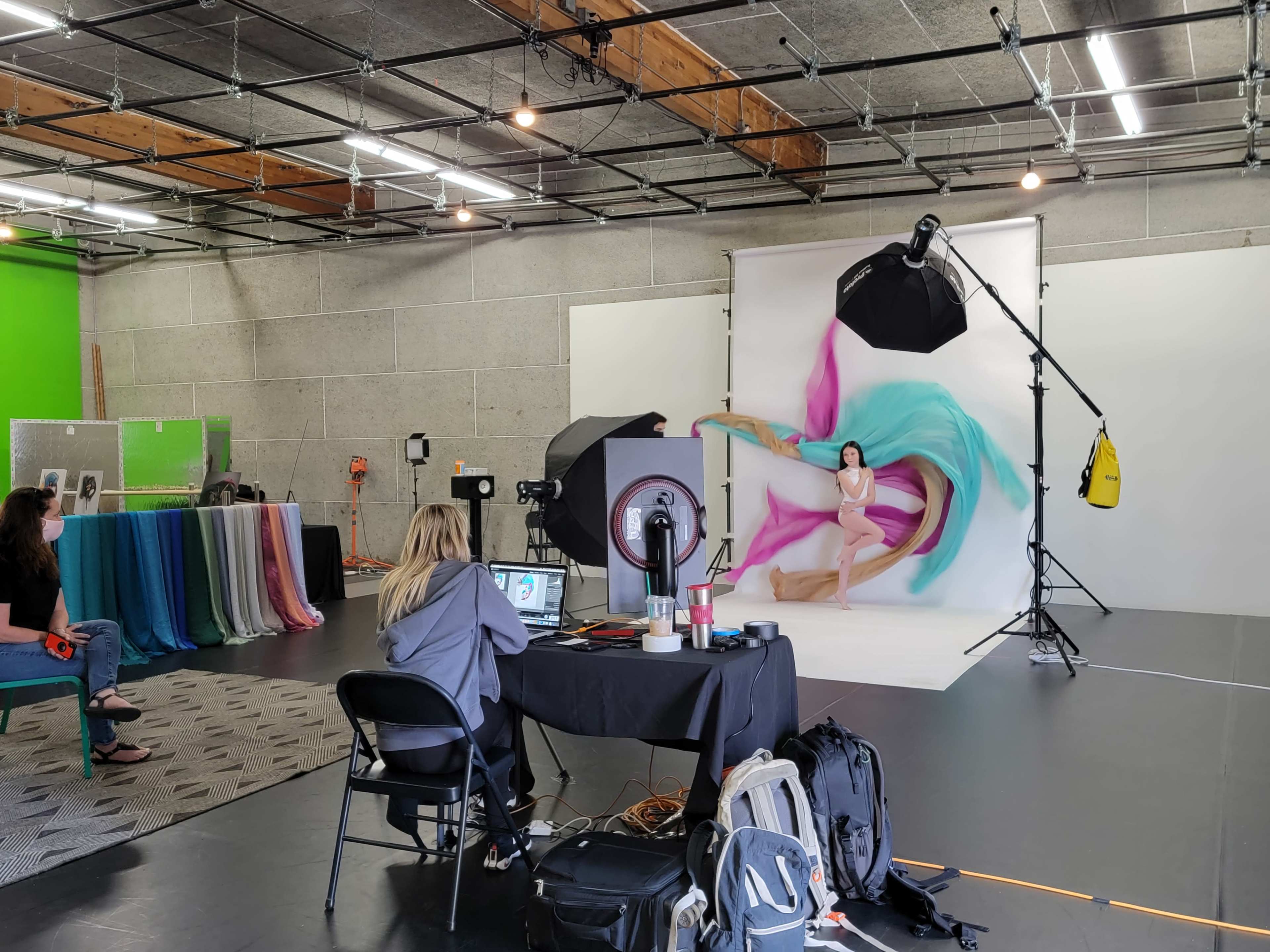 A photographer sets up a photo shoot with a model against a colorful backdrop in a studio with various fabric drapes and lighting equipment.