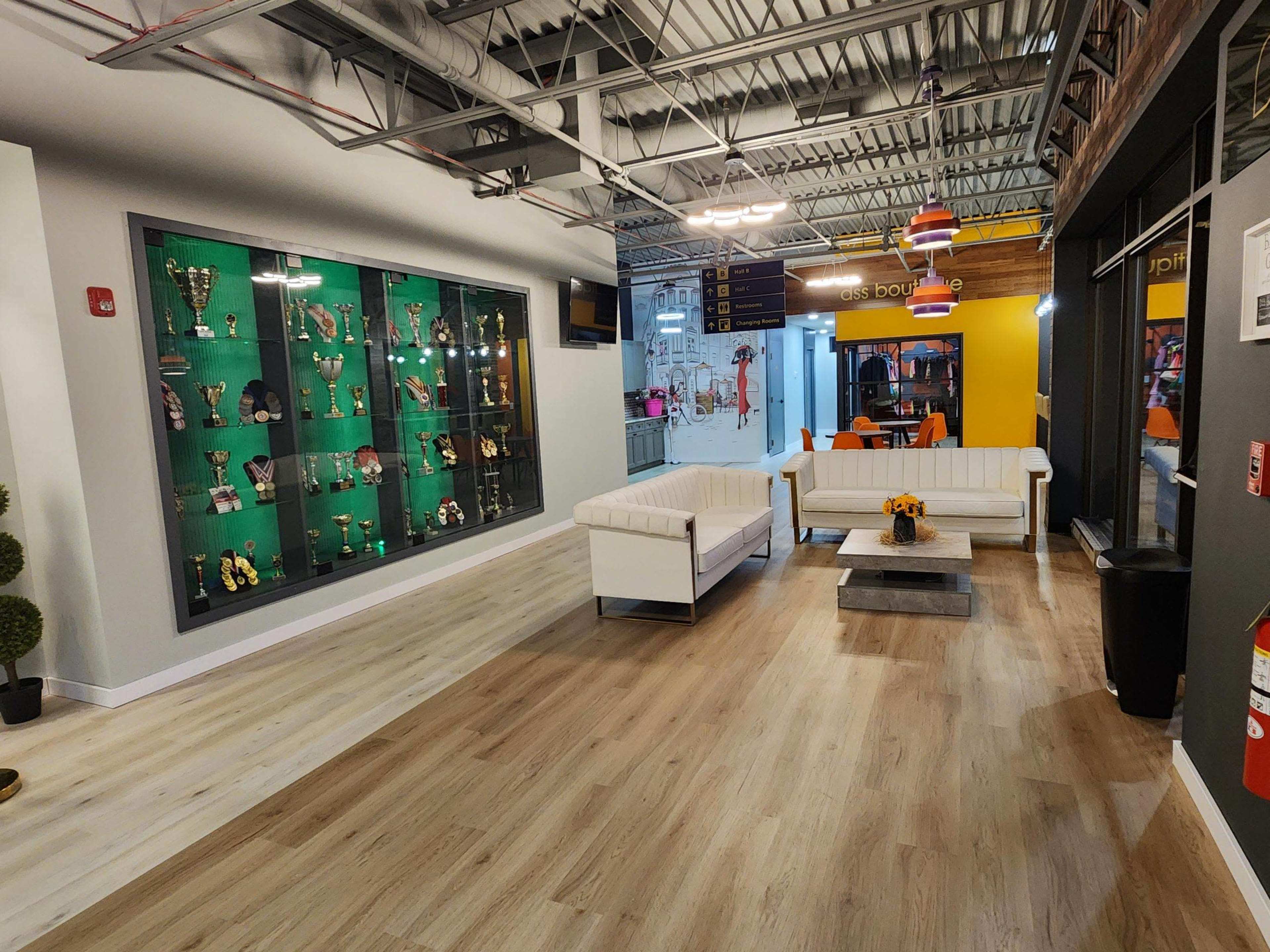 The image shows a modern lobby area featuring white sofas, a coffee table, and a display wall filled with trophies, alongside large windows and polished wooden flooring.