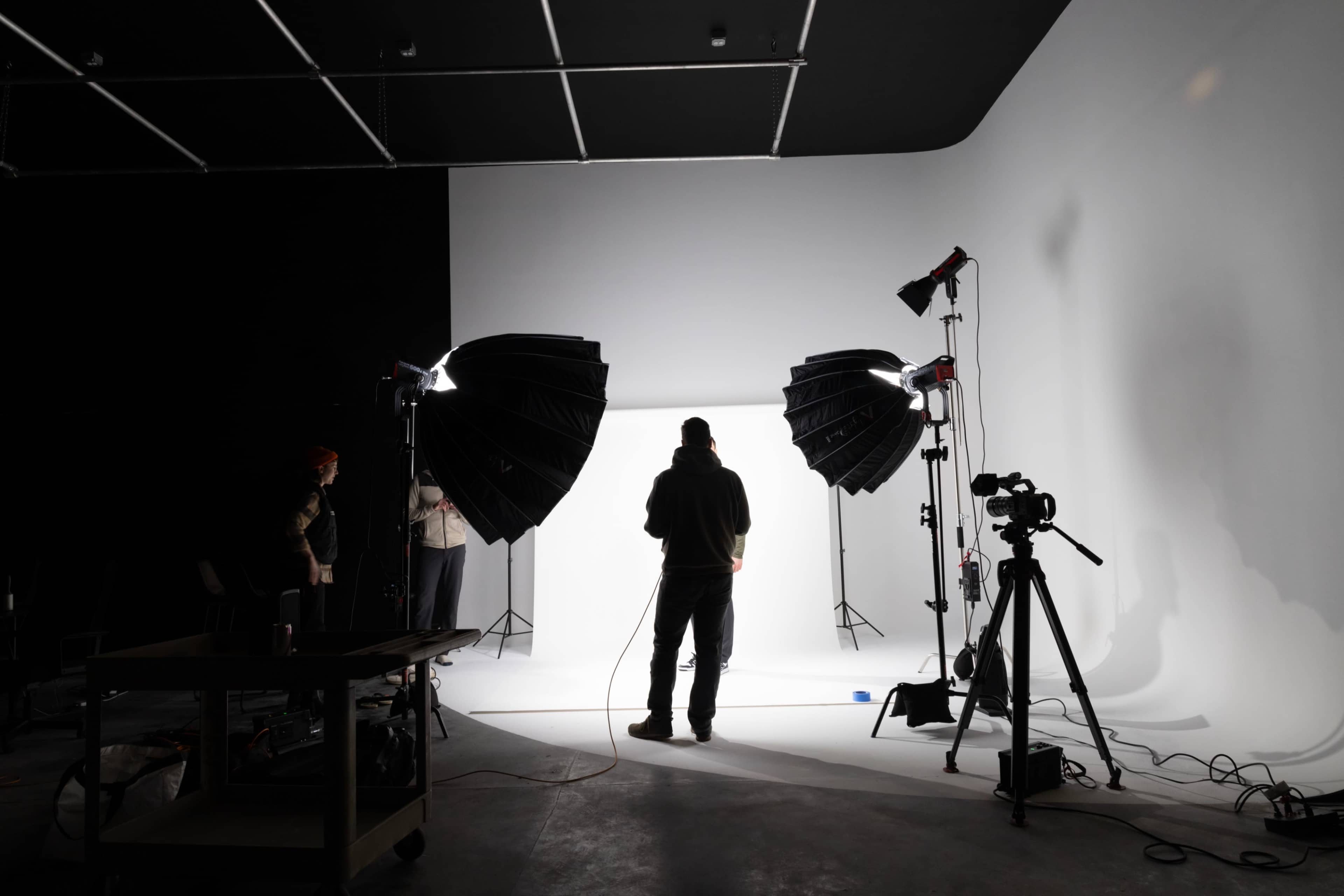 A person stands in front of a backdrop in a photography studio, with softbox lights set up on either side and a camera on a tripod nearby.