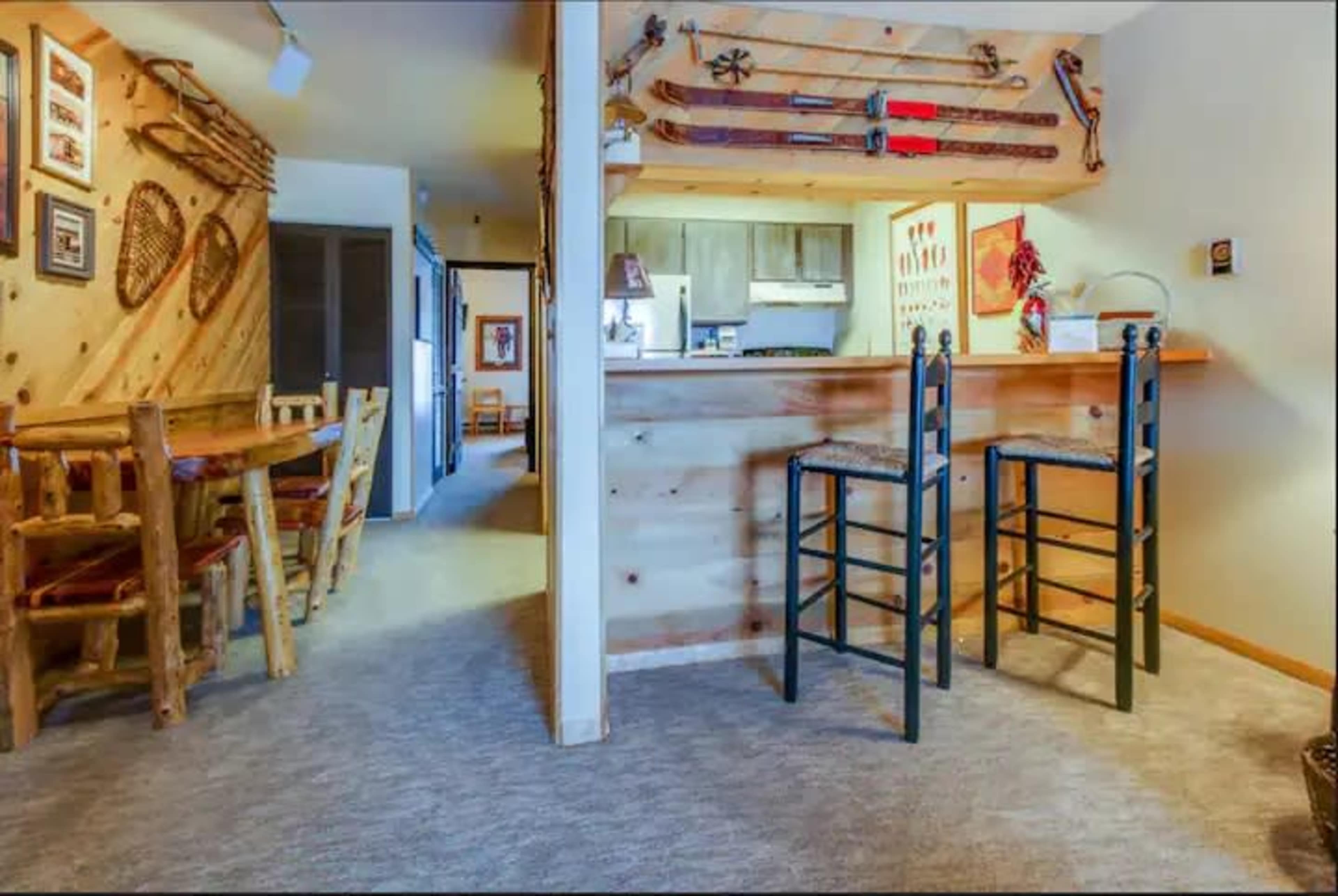 The image shows a cozy interior space with wooden walls, a small kitchen area, a bar with two black stools, and rustic decor including skis and snowshoes on display.