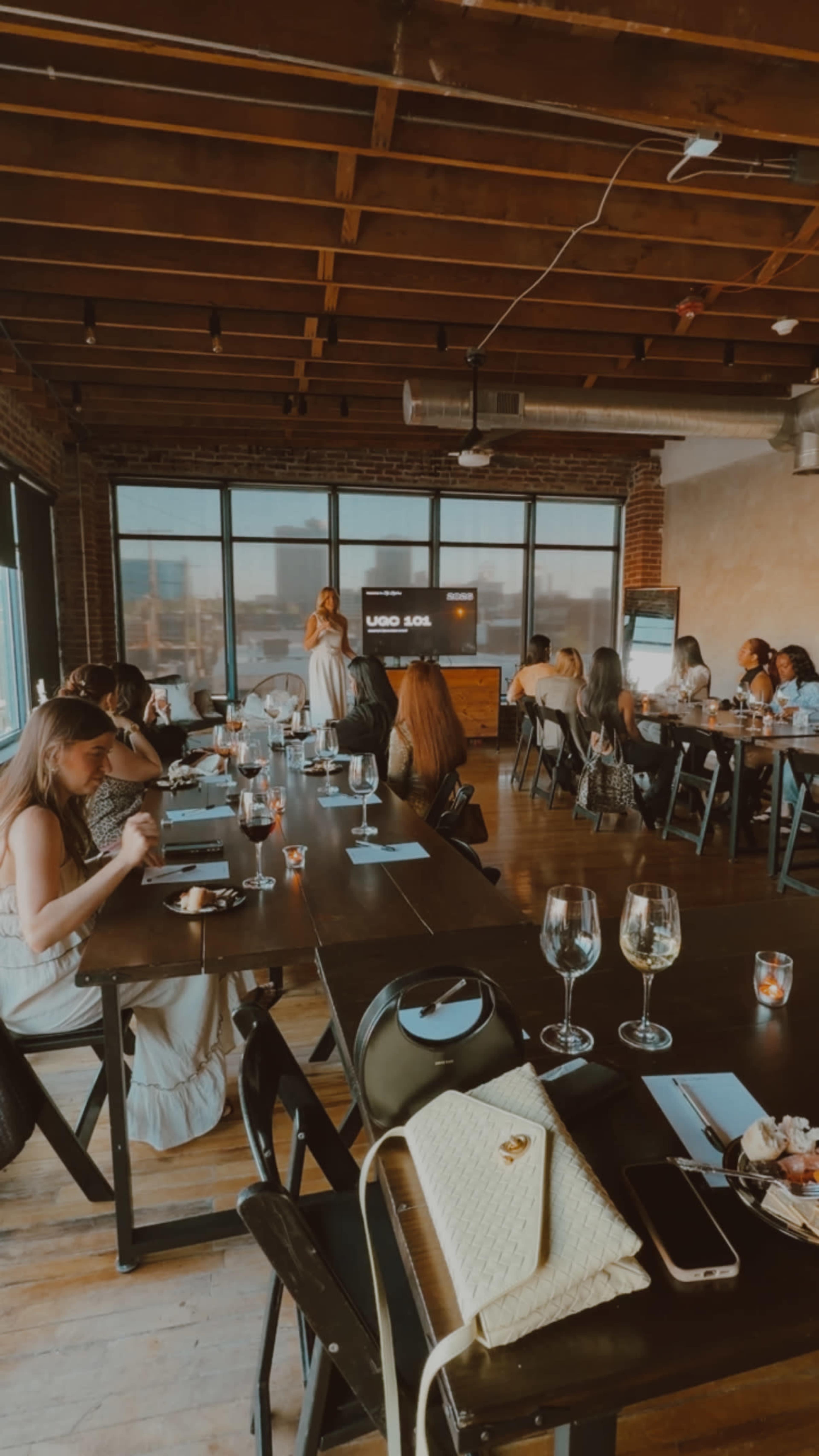 A group of people is seated at tables in a well-lit restaurant, some engaged in conversation while others are looking at their phones or enjoying their meals.