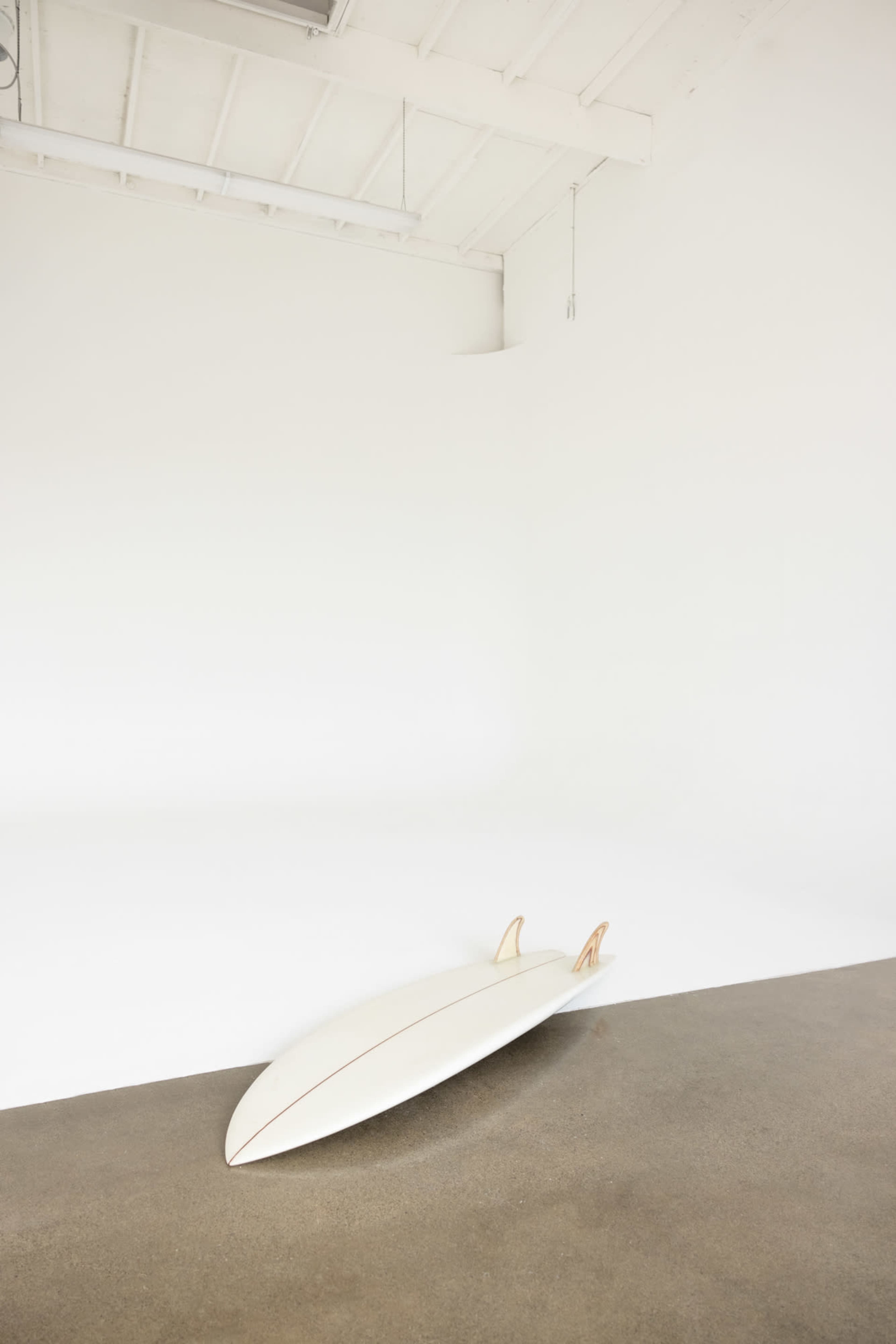 A surfboard leans against the wall of a minimalist, empty room with a concrete floor and white walls.