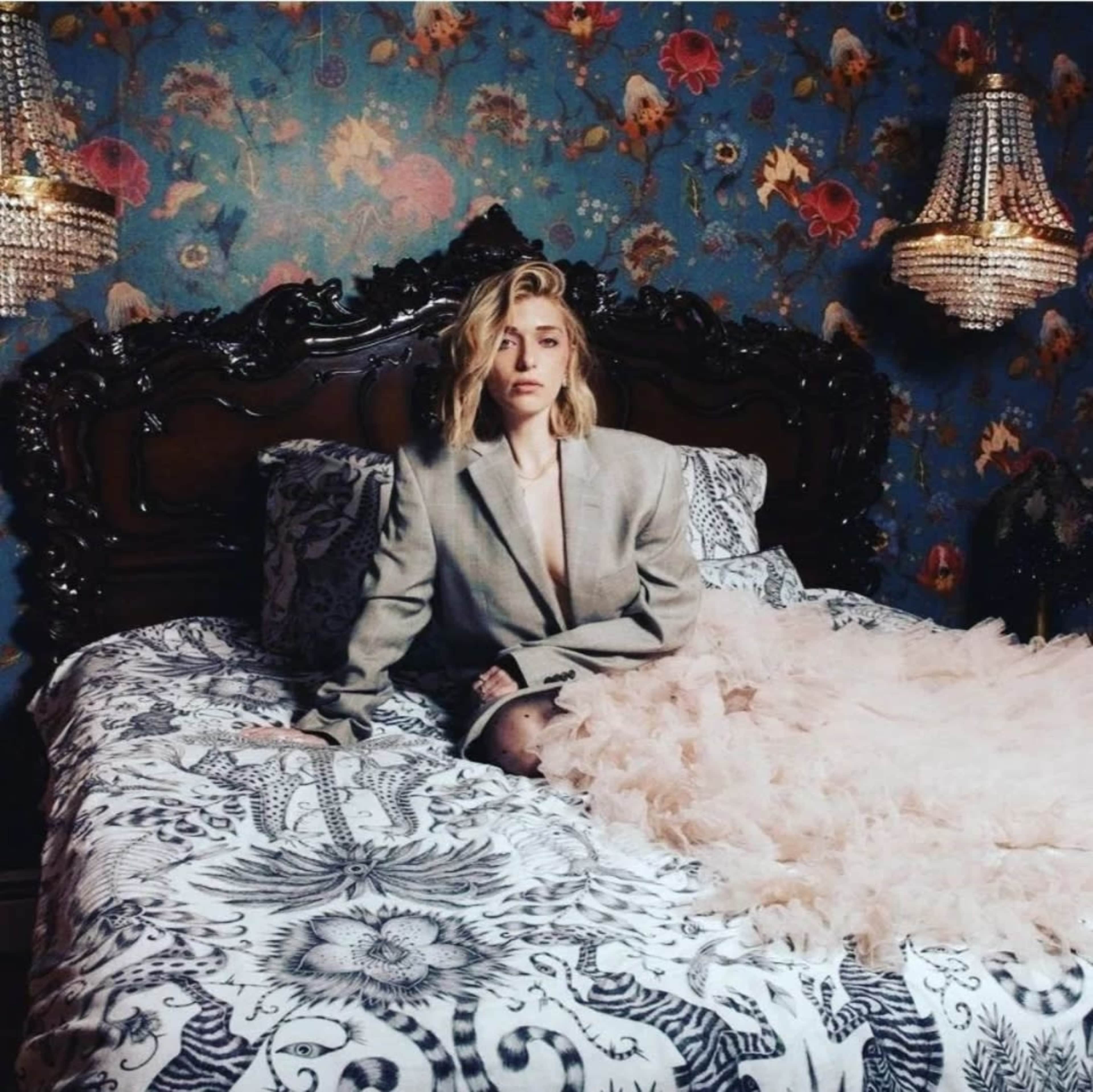 A woman in a beige blazer sits on a bed with ornate bedding and a floral wallpaper backdrop, surrounded by crystal chandeliers.