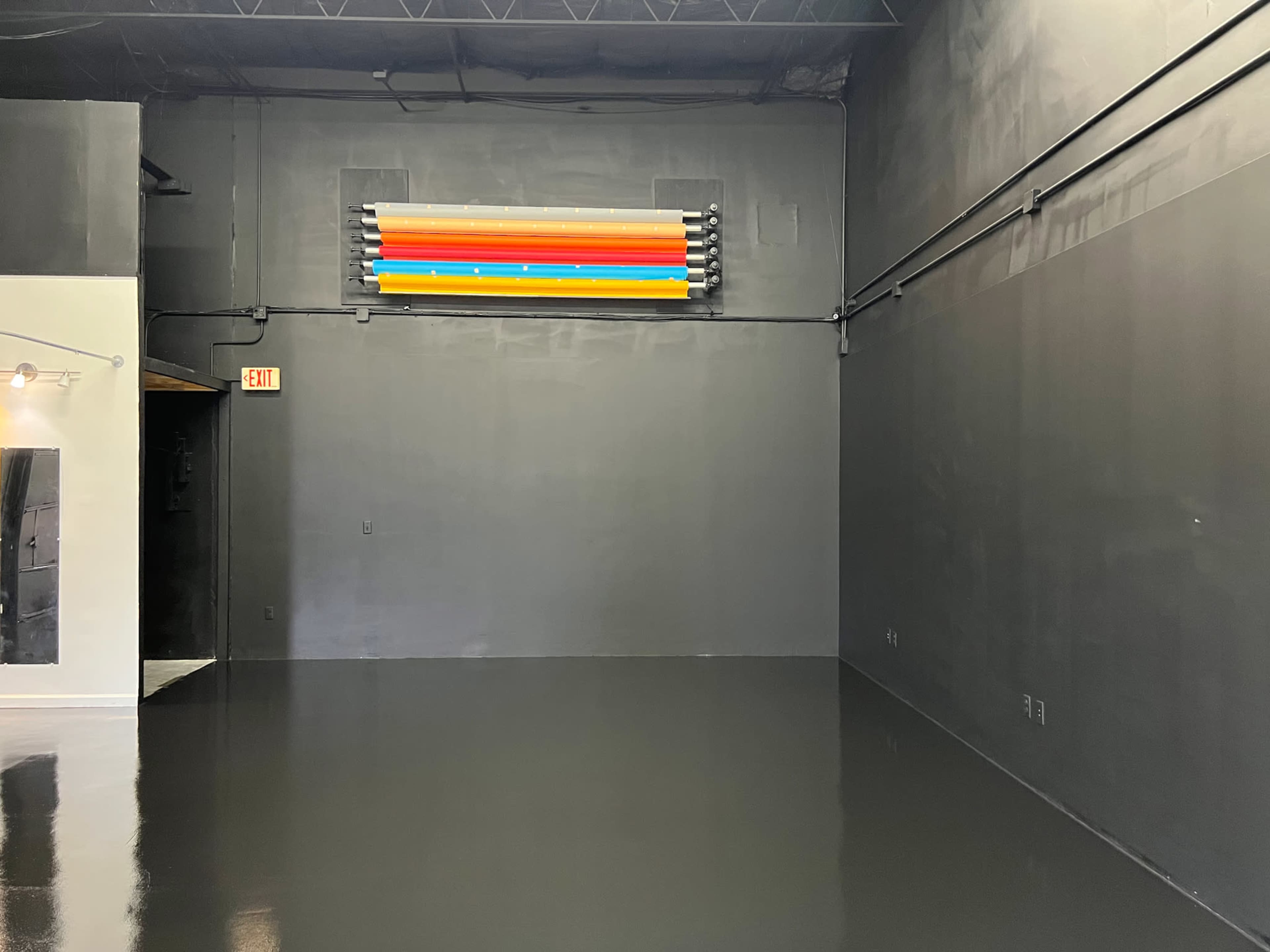 A sparse room with a glossy dark floor and a wall displaying colorful fluorescent light tubes.
