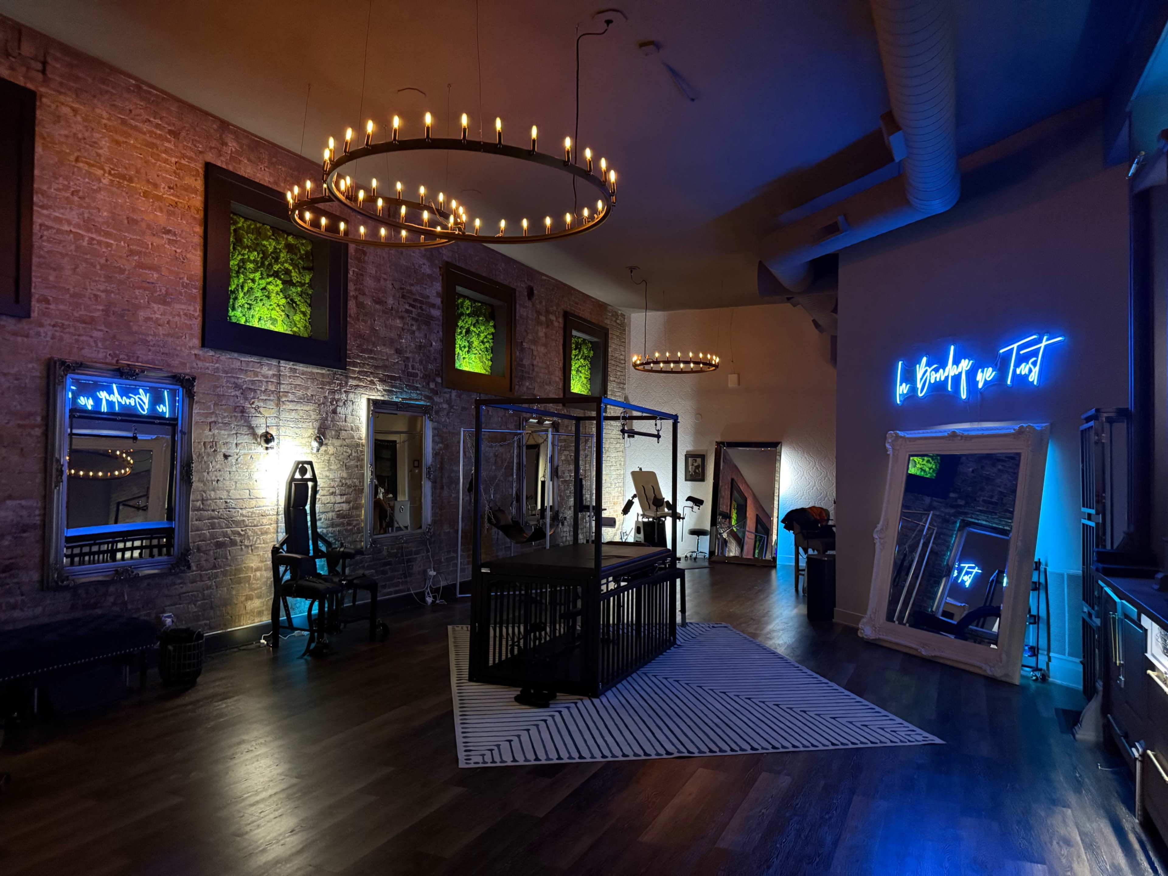 The image shows a spacious, well-lit room with exposed brick walls, featuring modern decor, mirrors, and a neon sign, alongside fitness equipment and decorative chandeliers.