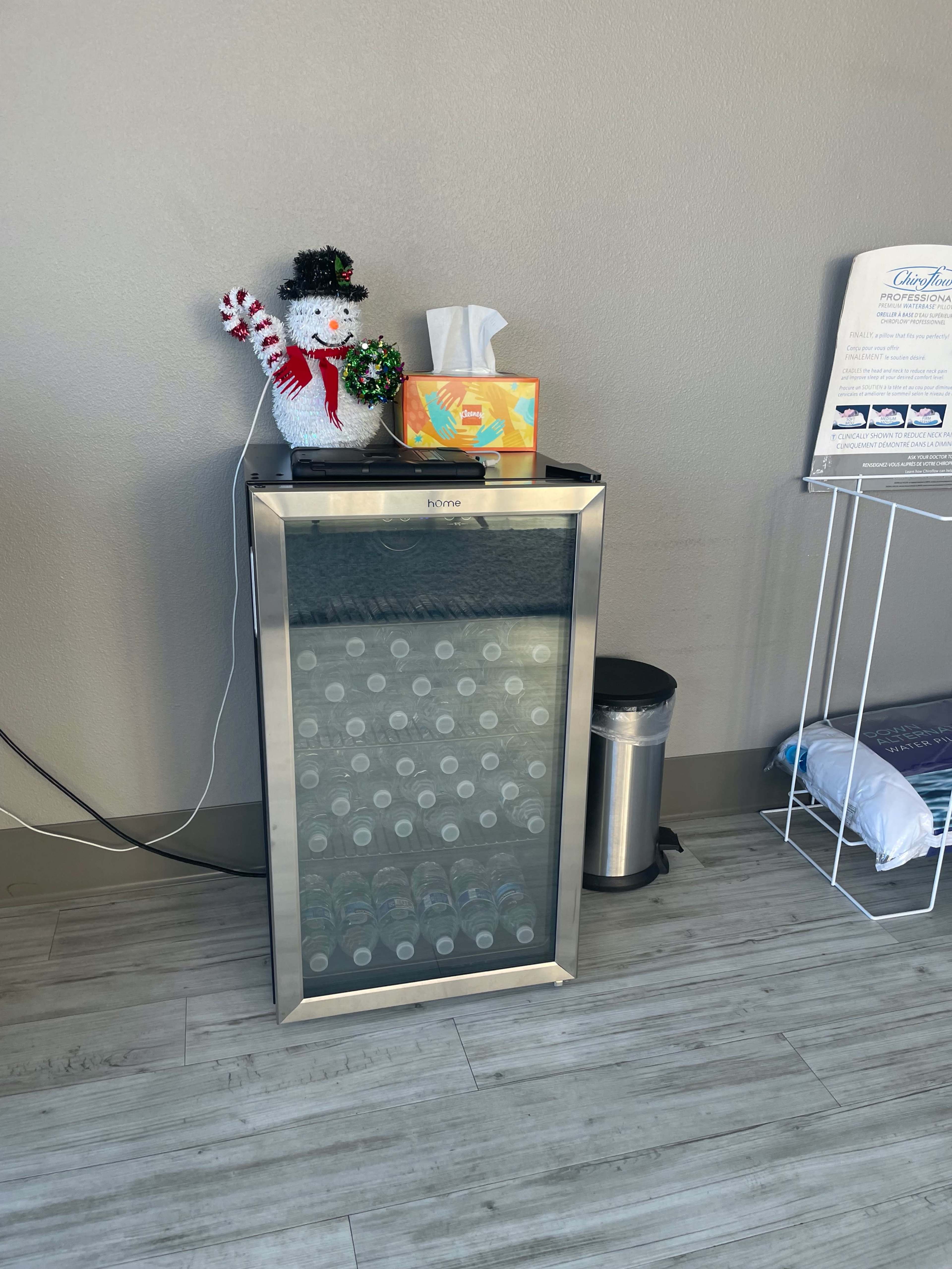 A small beverage cooler sits against a wall, topped with a snowman decoration, a box of tissues, and a colorful tissue box, next to a trash can and a shelf with supplies.