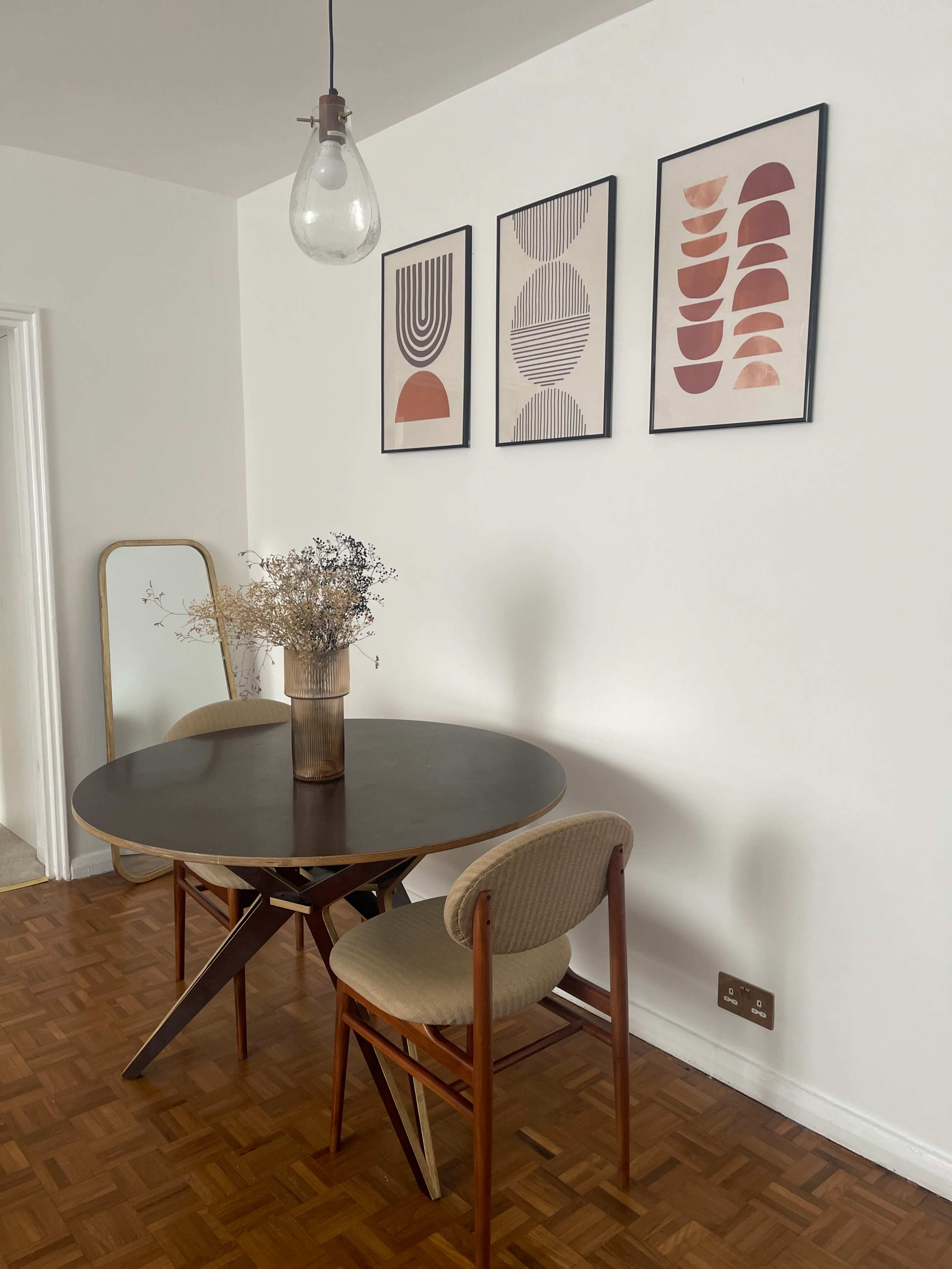 A round dining table with two chairs is positioned near a wall adorned with three framed artworks, while a vase with dried flowers sits in the center of the table.