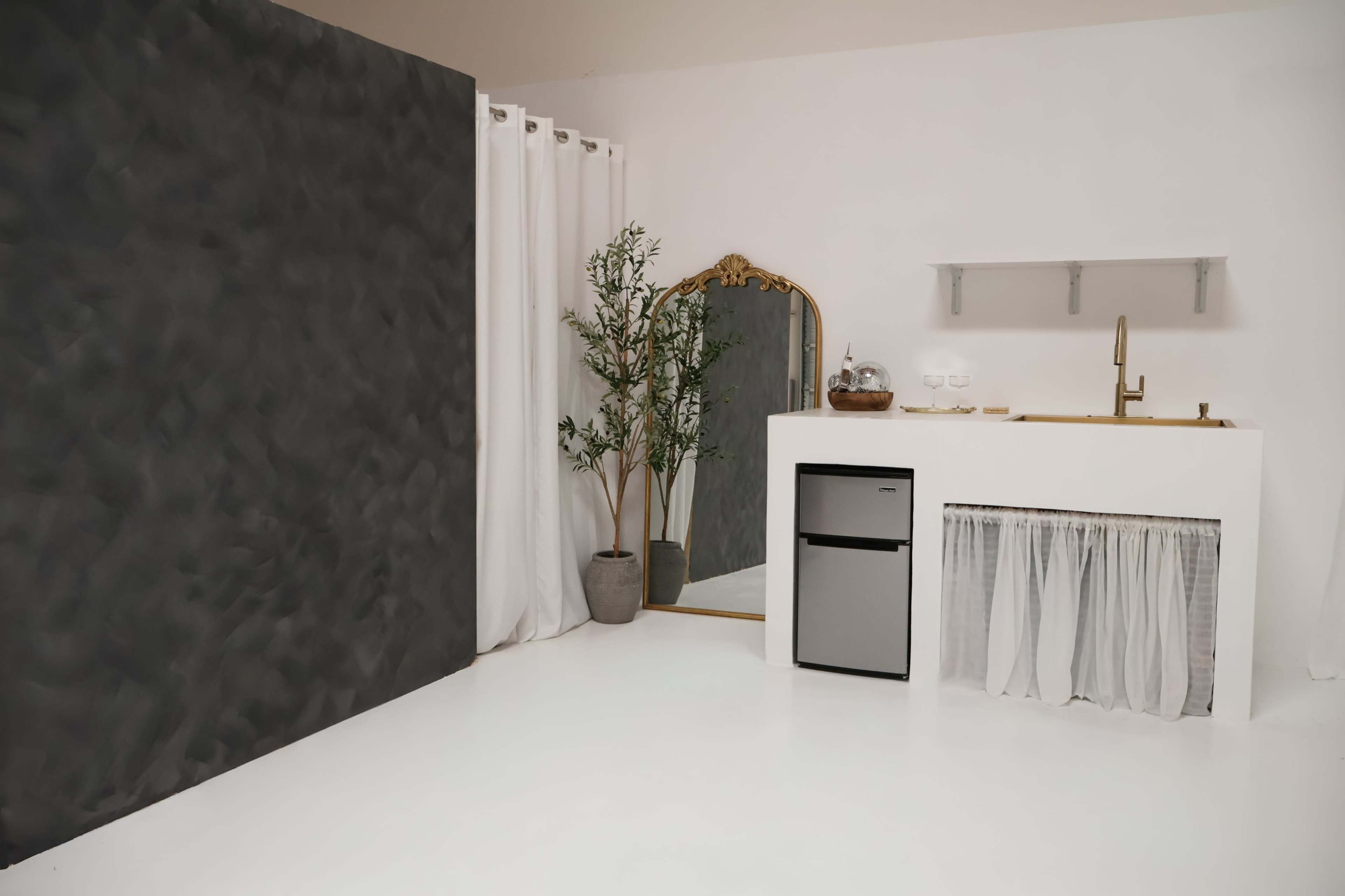 The image shows a minimalist interior space featuring a gray wall, a small kitchen area with a refrigerator, and a large mirror framed in gold, accompanied by a decorative plant in a pot.