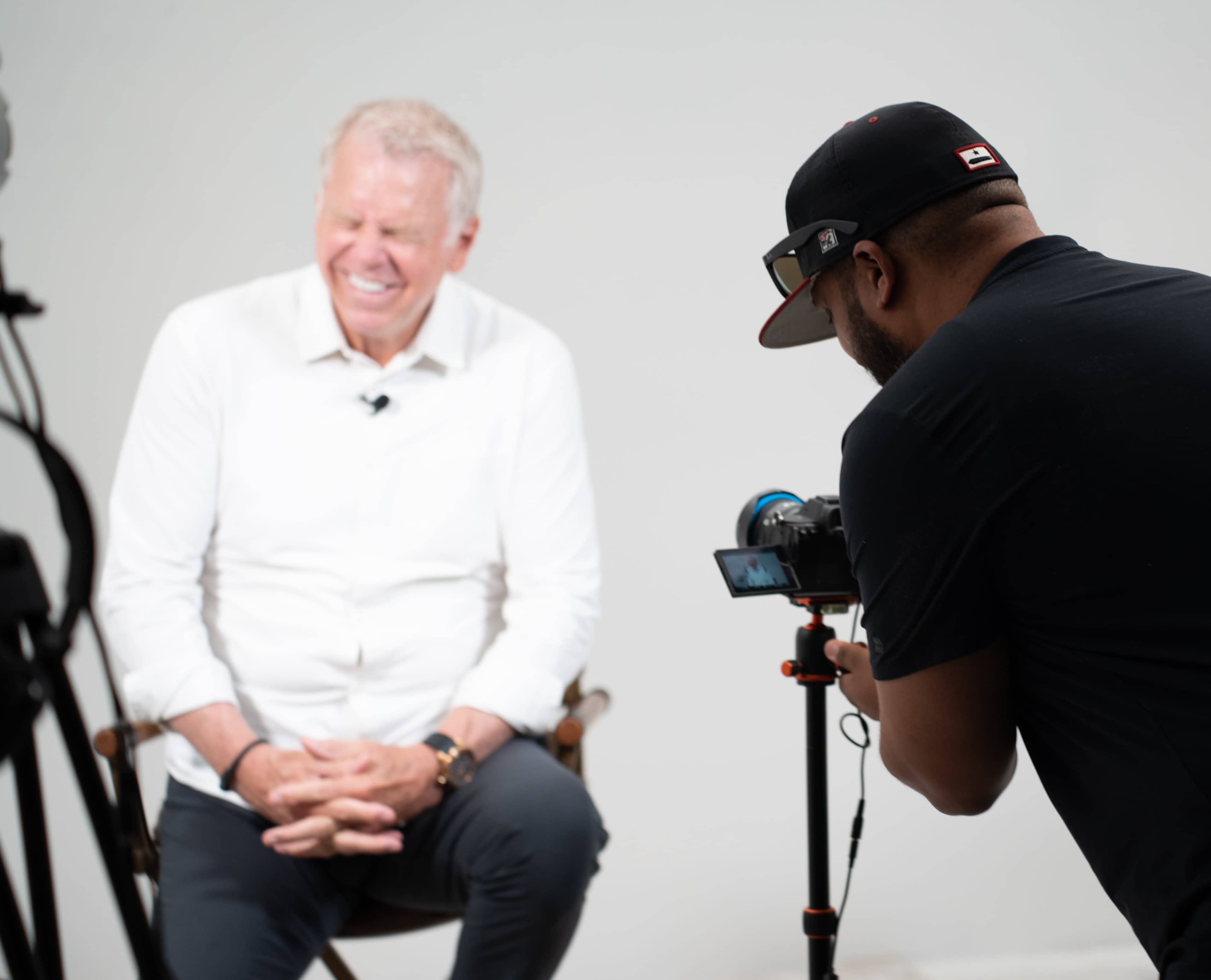 A light-haired man in a white shirt sits on a chair while another man with a camera prepares to take his photo.