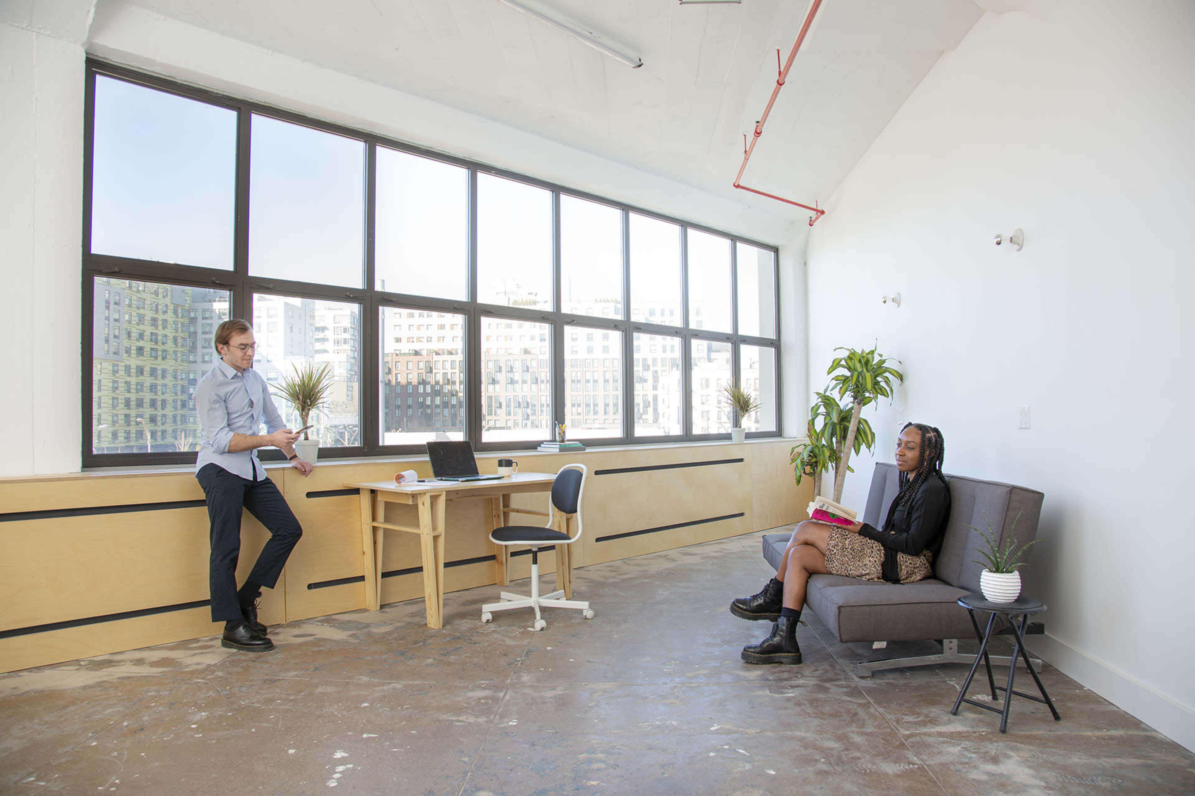 A man stands near a large window in a modern office space while a woman sits on a sofa reading a book.