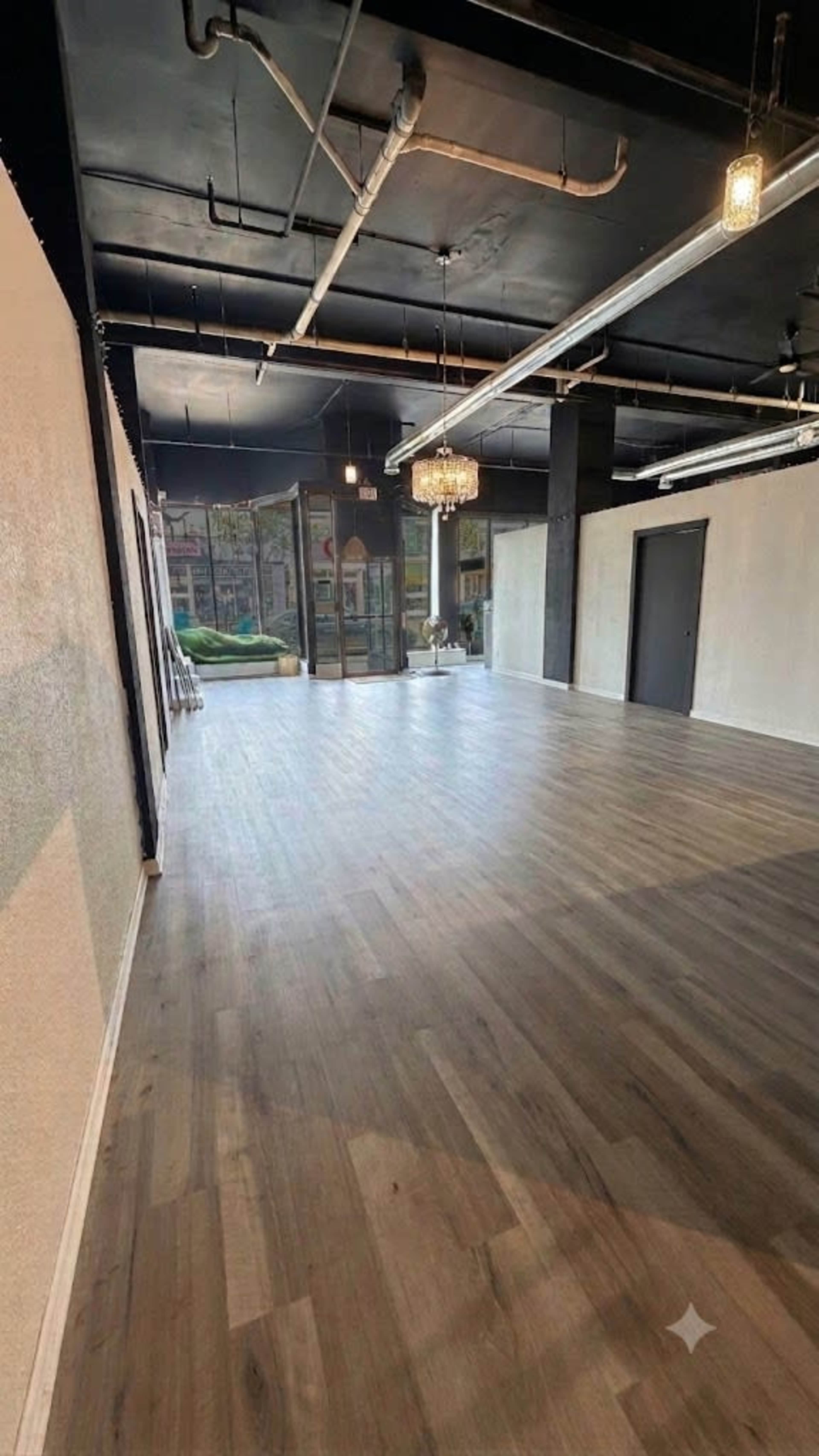 The image shows a spacious interior with wooden flooring, large windows, and minimal furnishings, leading out to an outdoor area.