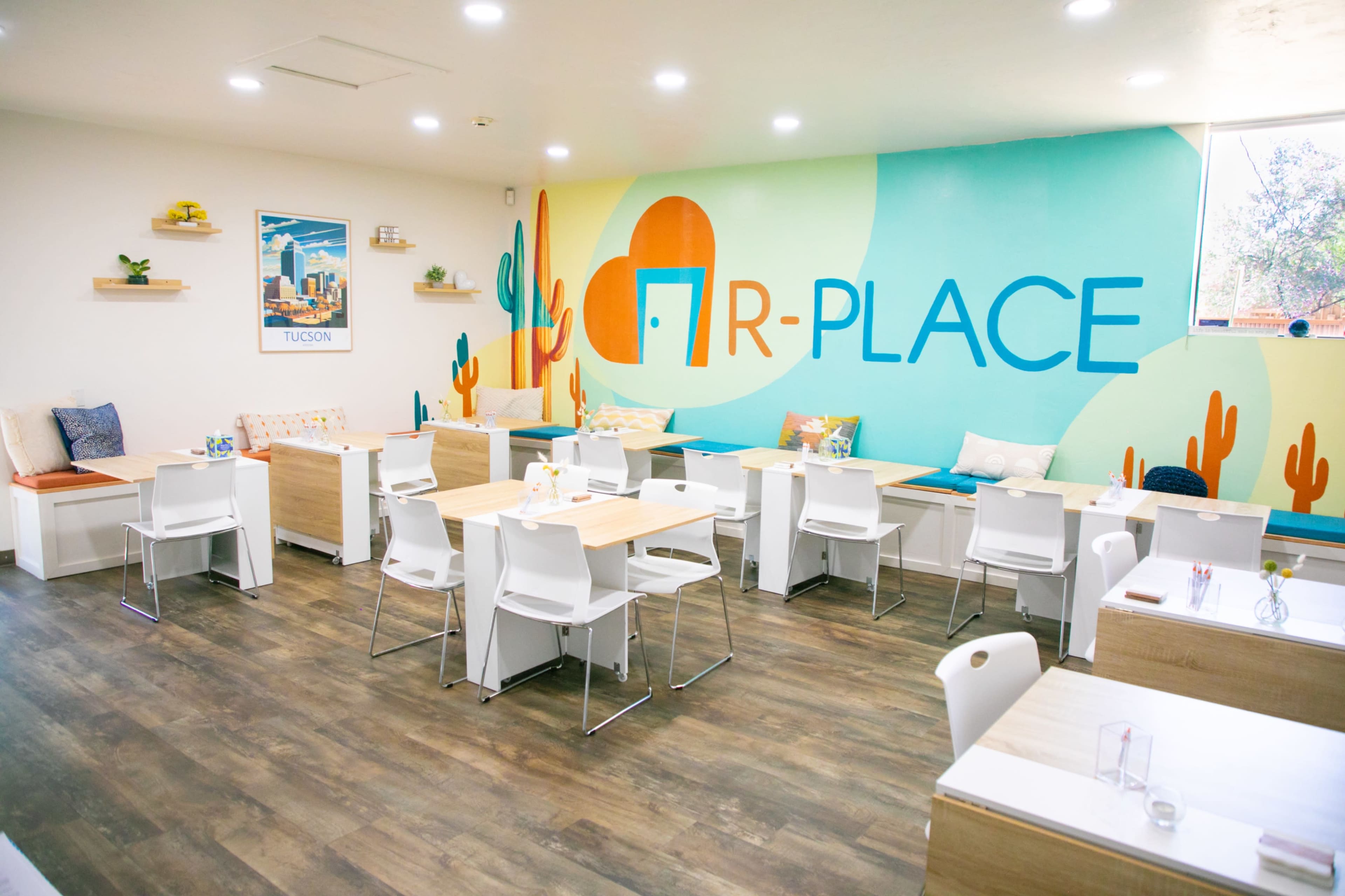 A brightly colored café interior with light wooden tables, white chairs, and a mural featuring cacti and the word "PLACE" on the wall.