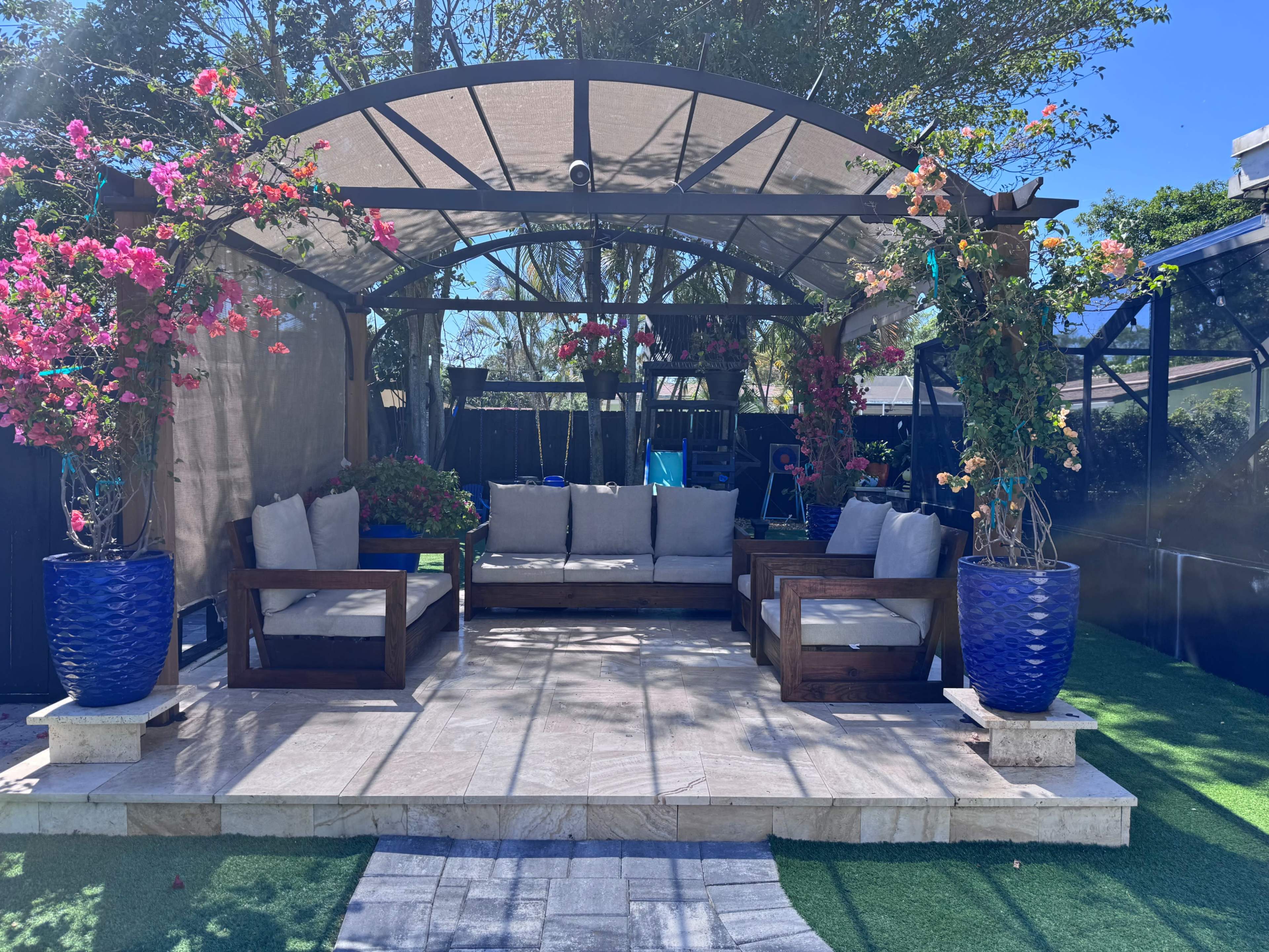 A covered outdoor seating area features cushioned sofas and vibrant potted plants on a stone patio.