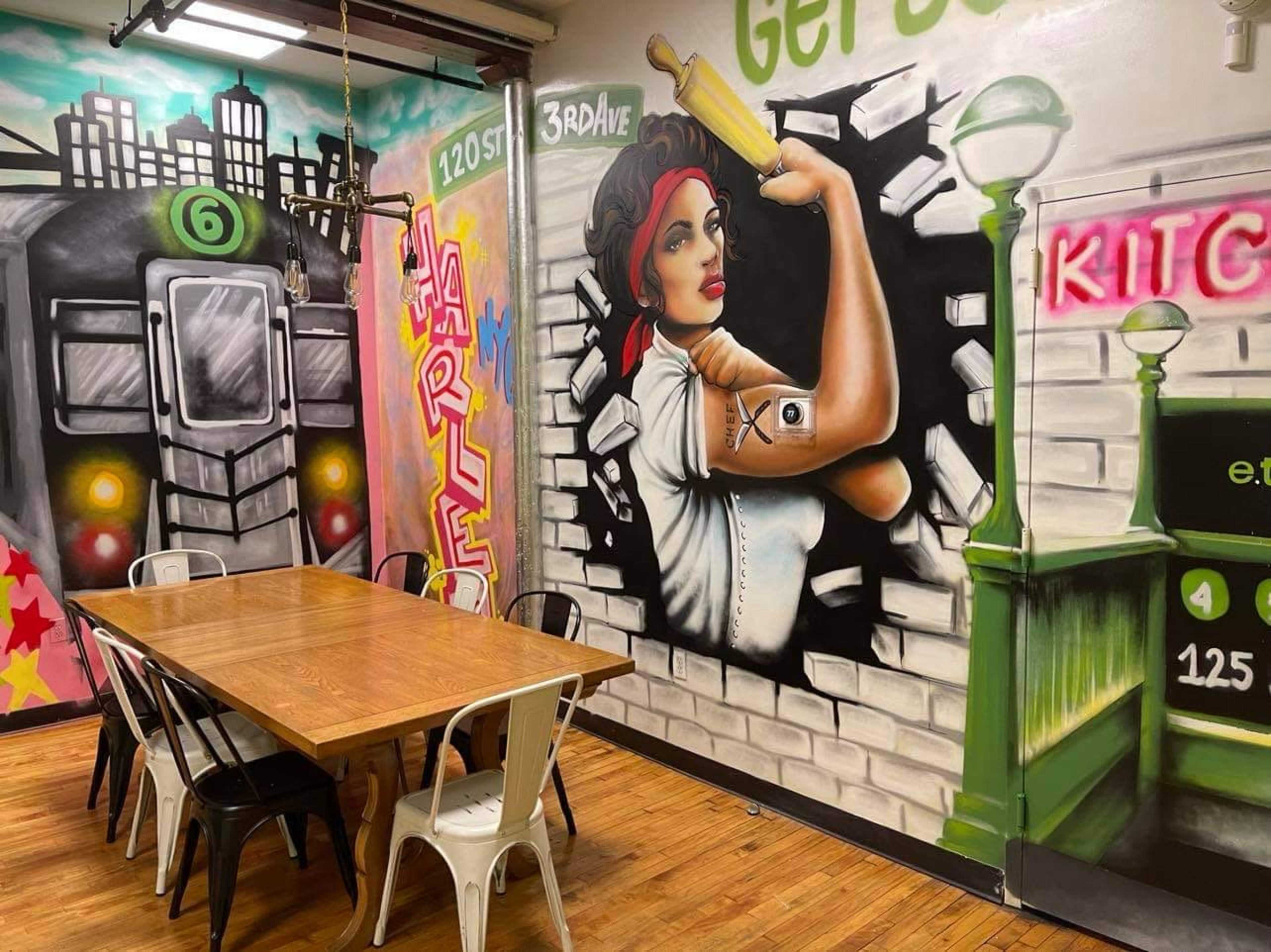 A dining area with a wooden table and metal chairs, surrounded by vibrant murals depicting a woman breaking through a wall and a city skyline.