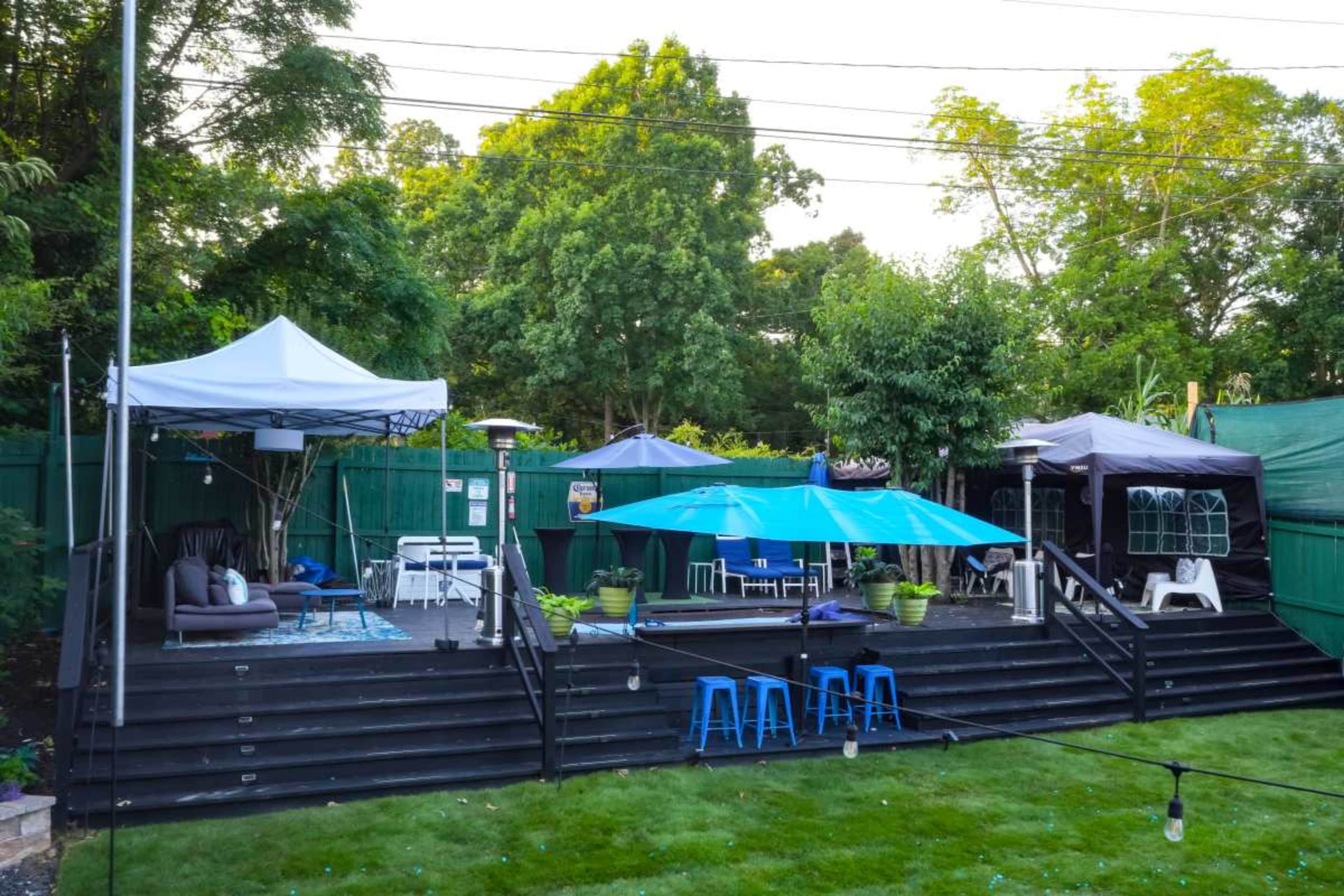 The image depicts a backyard with wooden steps leading to an outdoor seating area, featuring various tents, tables, and chairs surrounded by greenery.