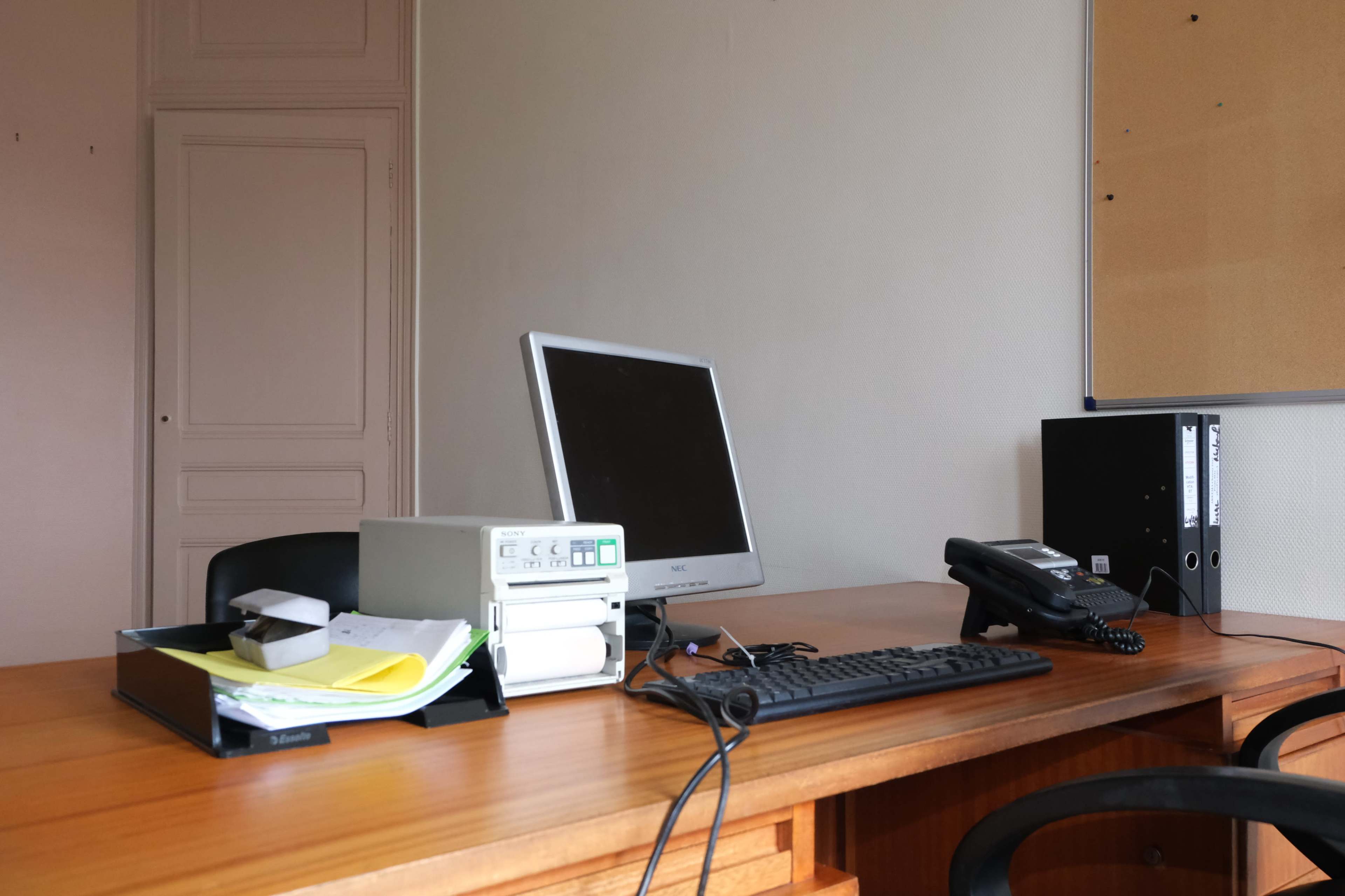 A simple office setup featuring a desktop computer, a phone, and assorted paperwork on a wooden desk.